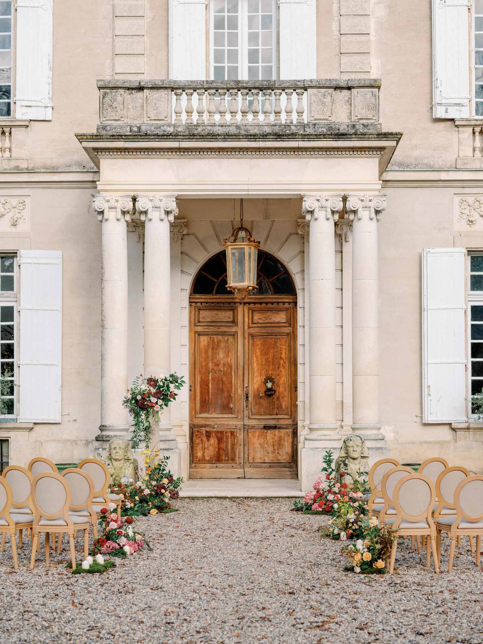 Outdoor ceremony with wooden chairs and coral and blush floral arrangements before a stone mansion with Corinthian columns