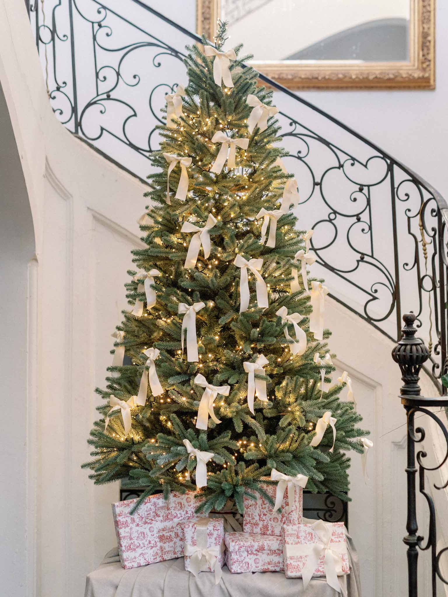 Decorated Christmas tree with cream ribbon bows, gold lights, and red toile wrapped gifts on staircase