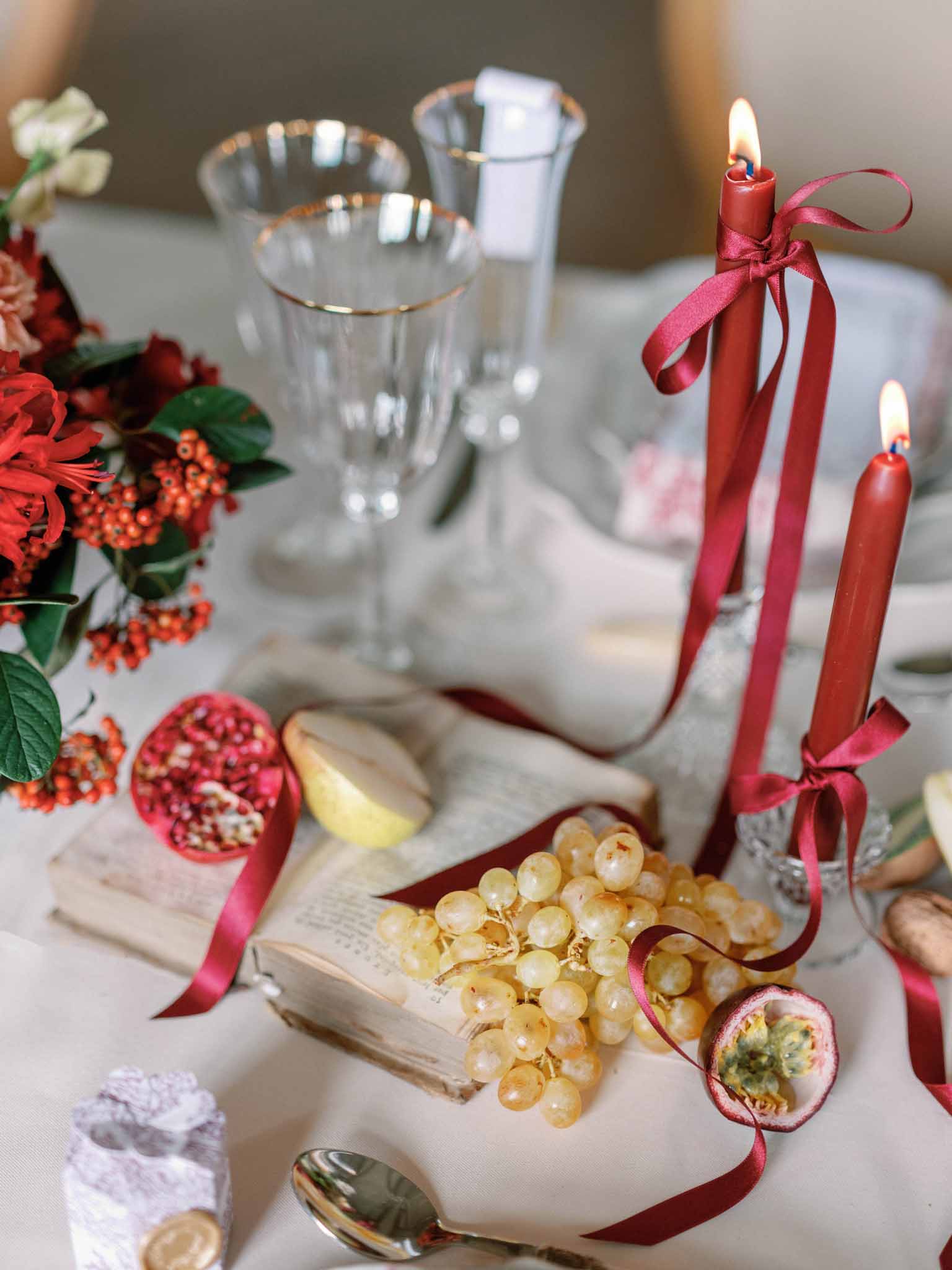 Reception table close-up with gold-rimmed crystal glasses, red tapered candles tied with pink ribbon, and fruit and floral styling