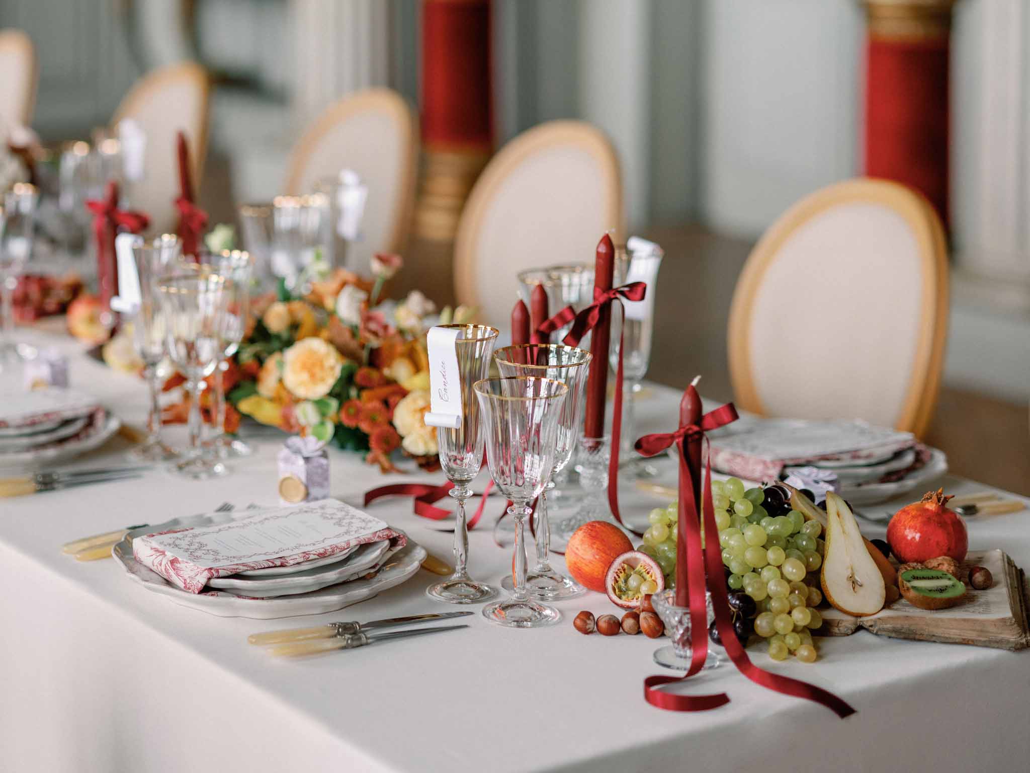 Reception table with stacked plates, burgundy candles, gold cutlery, and autumn floral runner with fresh fruits