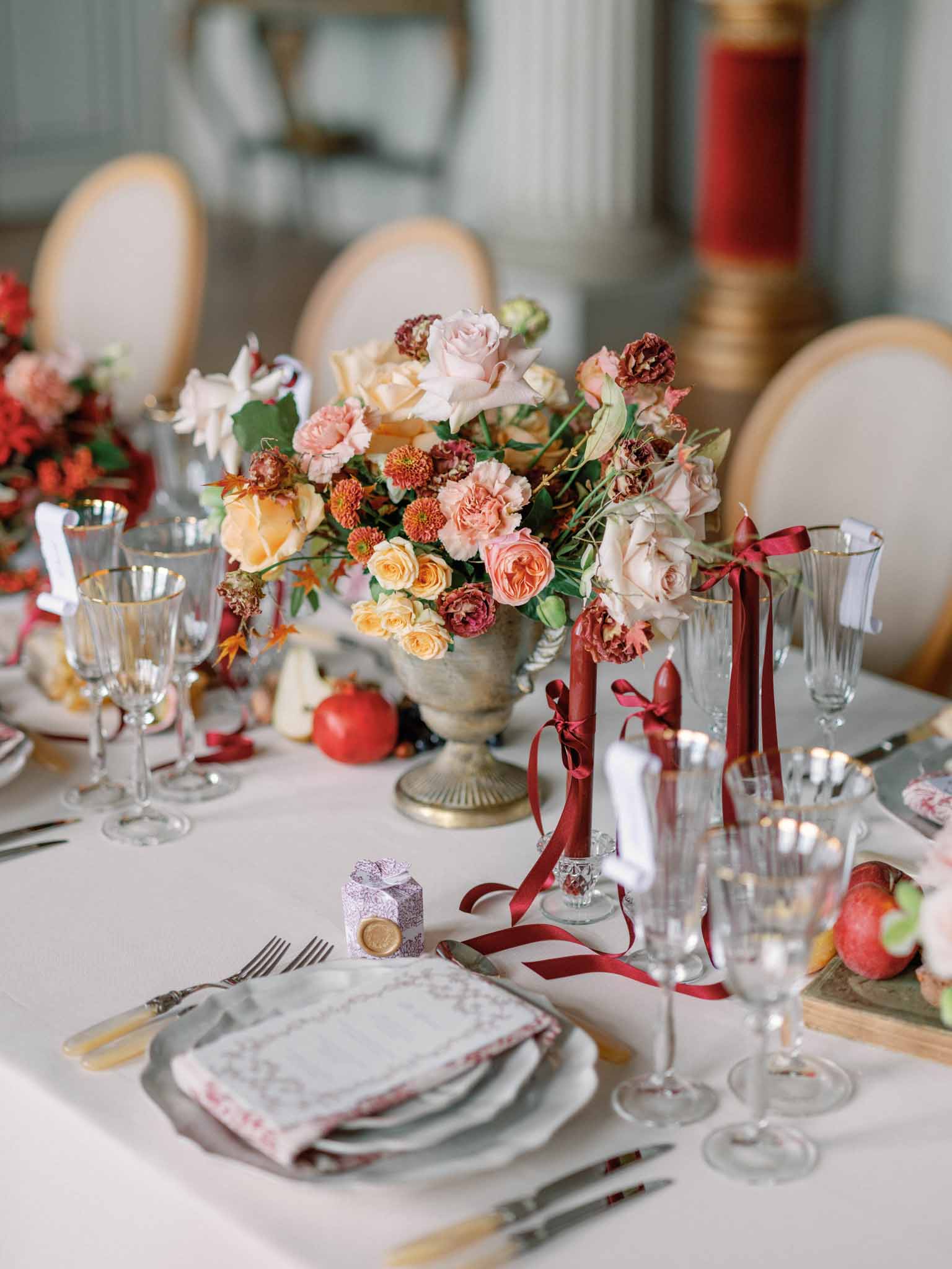 Close-up of reception table with peach garden roses, burgundy dahlias in urn, gold cutlery, and pomegranate accents