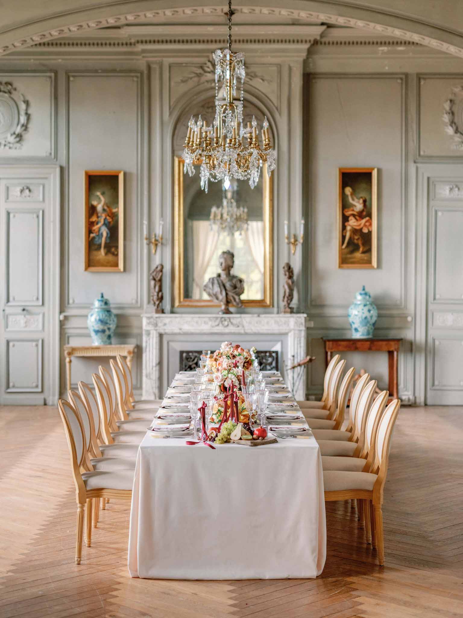 Long reception table in classical French ballroom with coral centerpieces, crystal chandelier, and gilt-framed paintings