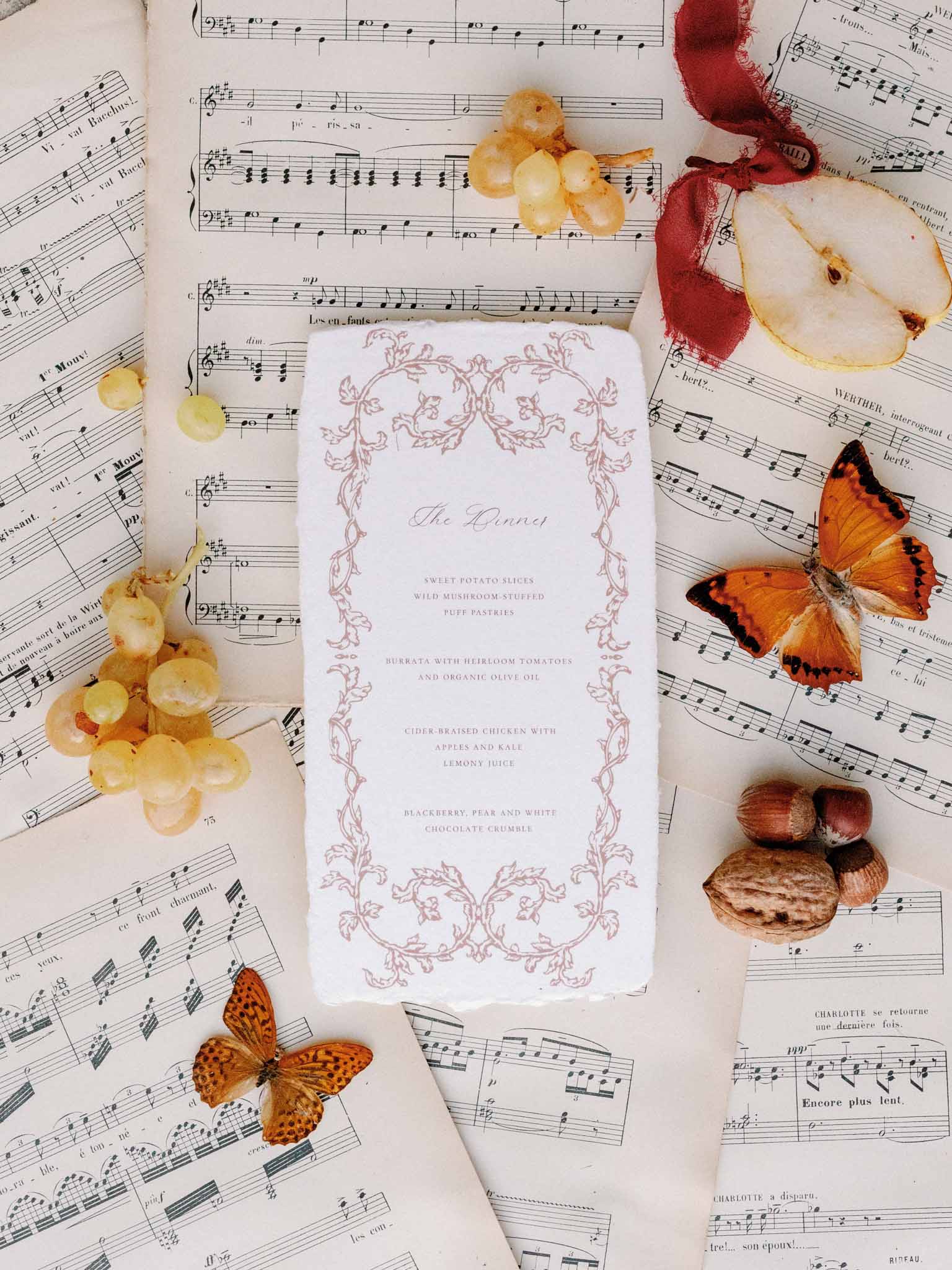 Flat lay of wedding dinner menu on vintage sheet music with dried orange butterflies and red ribbon accents