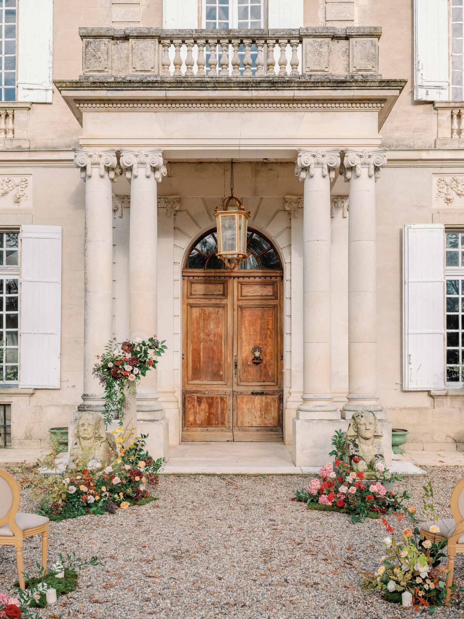 Neoclassical chateau entrance with Corinthian columns and coral floral arrangements flanking stone busts, gravel courtyard