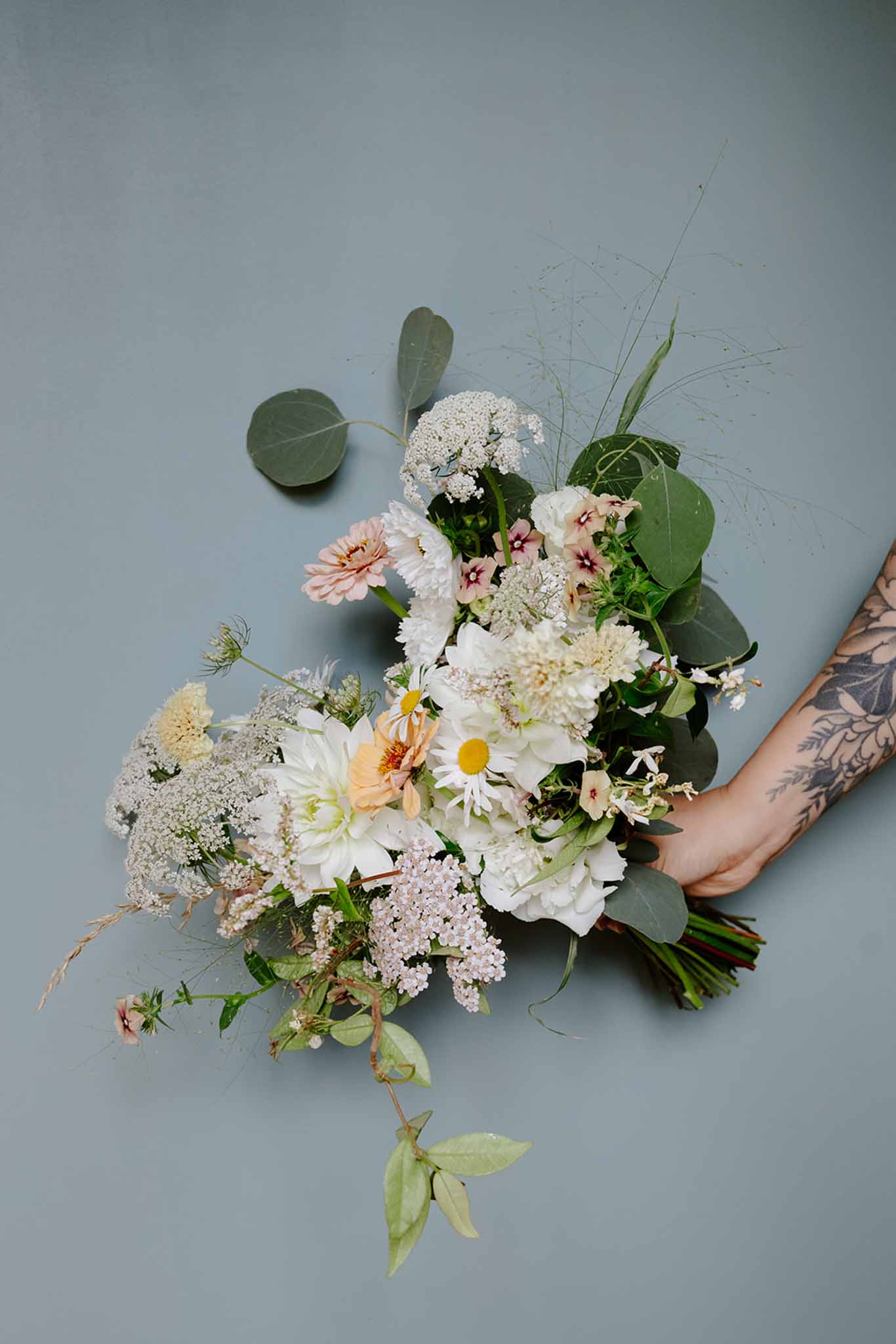 Bridal bouquet with white hydrangeas, blush dahlias, daisies, astilbe, and eucalyptus held by tattooed arm