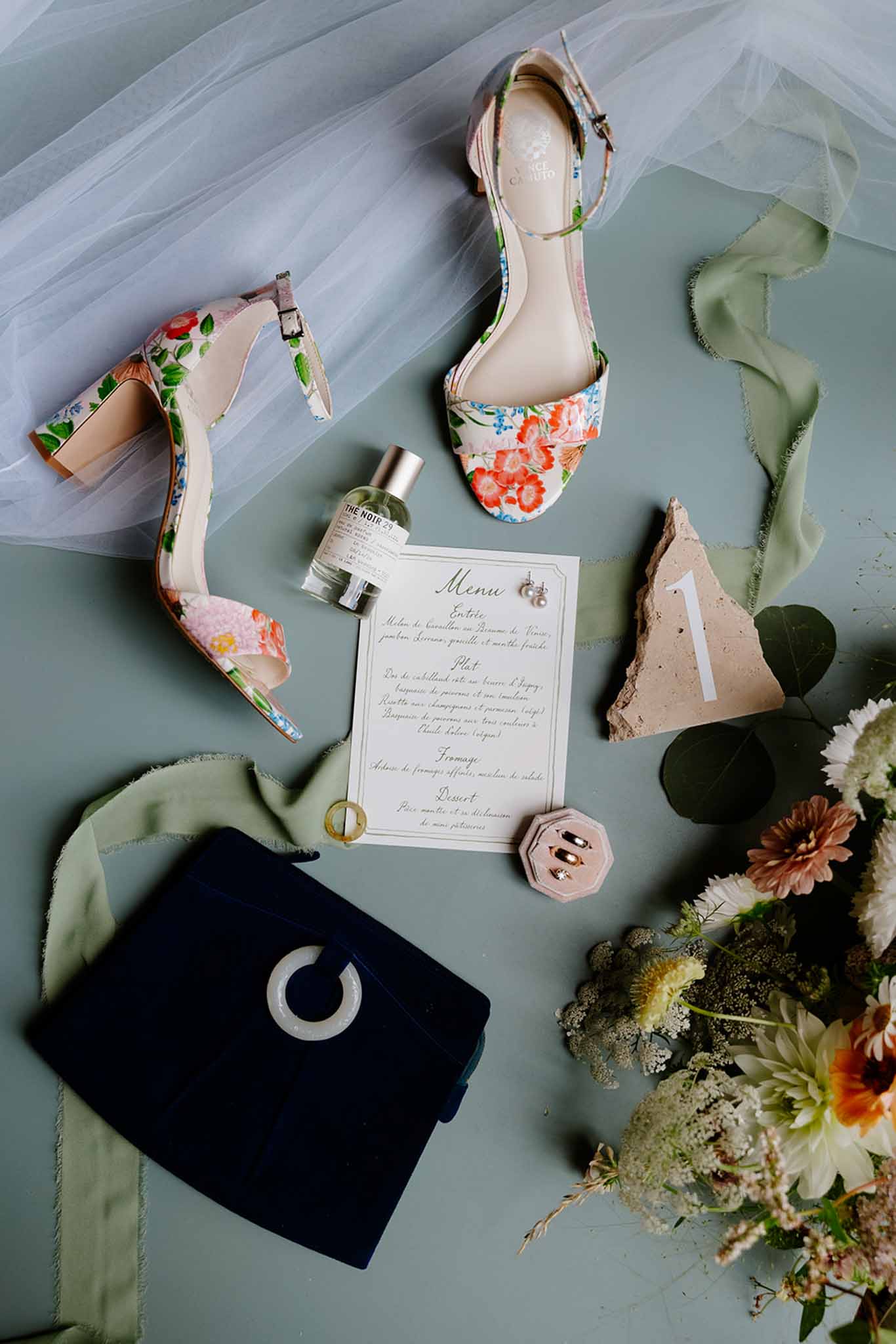 Flat lay on sage-green surface: floral-print shoes, calligraphy menu card, perfume, coral dahlias and a navy velvet clutch.