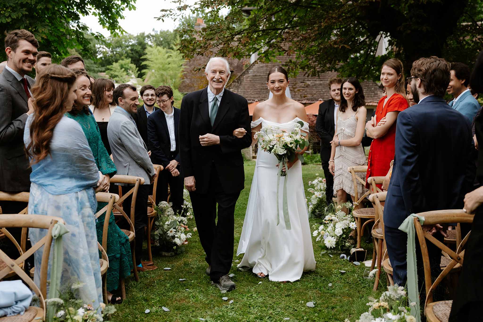 Bride in off-shoulder ivory gown walking aisle with elderly escort past cross-back chairs at stone manor