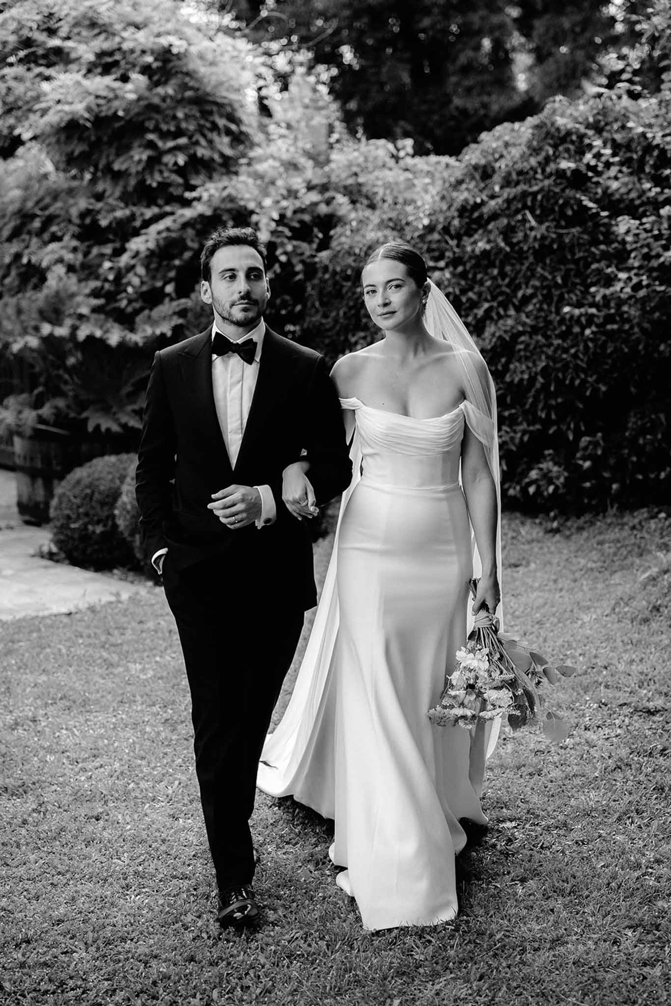 Black and white portrait of bride and groom walking together through a manicured garden, bride in off-shoulder gown with veil.