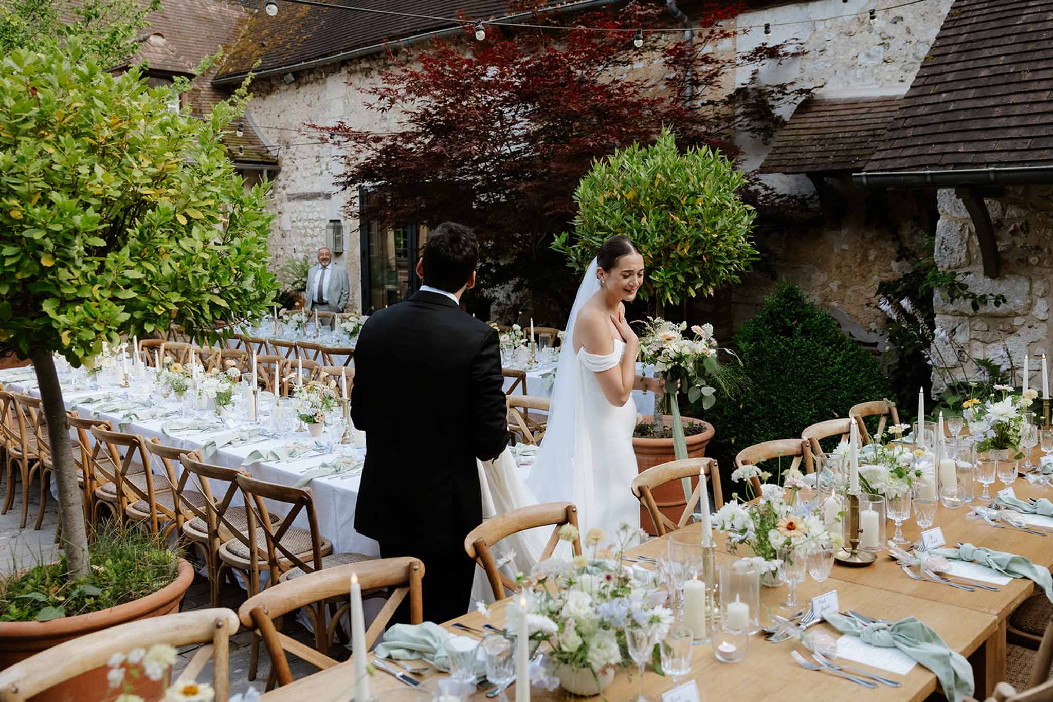 Bride and groom walking through outdoor courtyard reception with long wood tables, sage green napkins and white florals