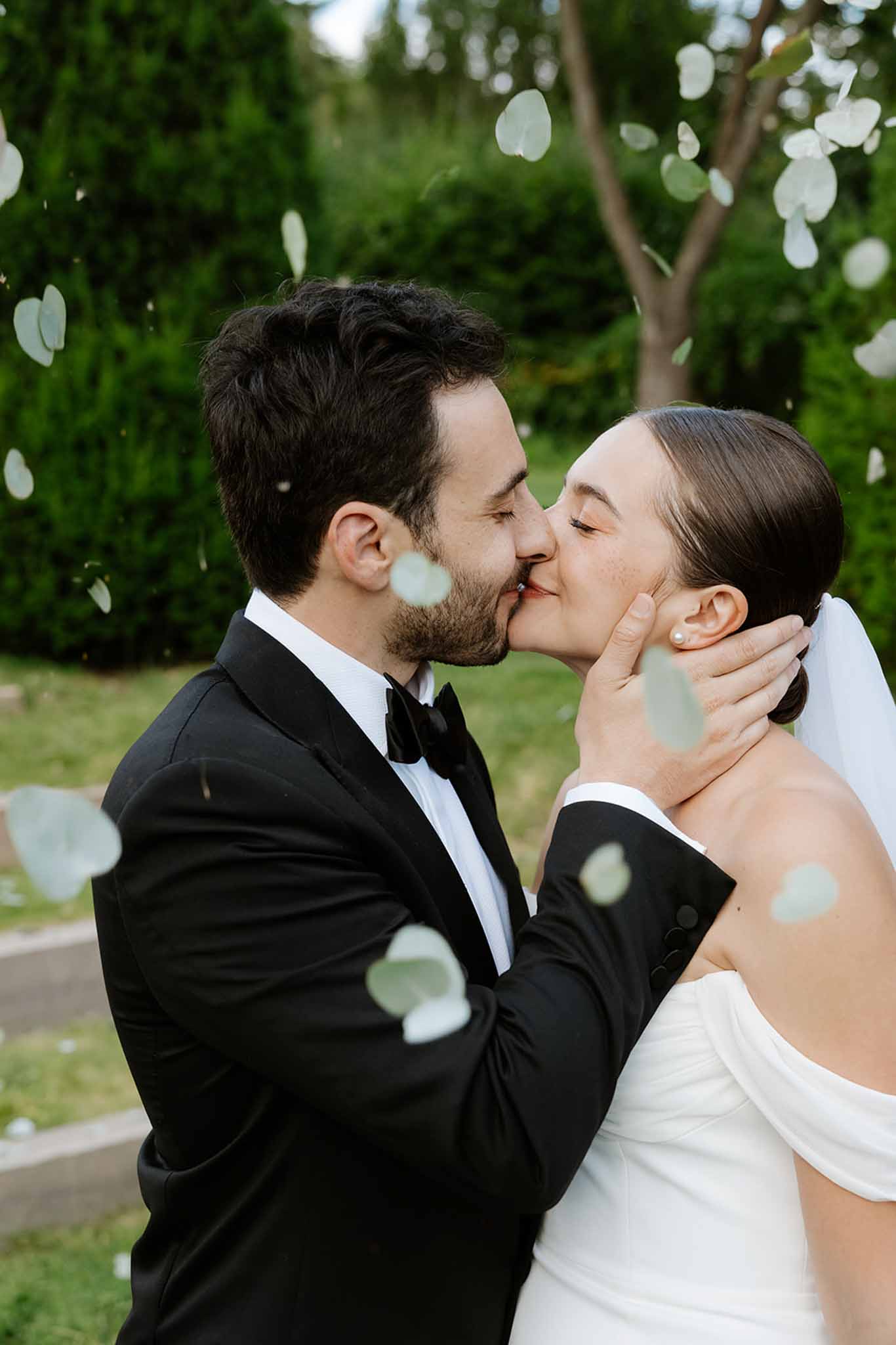 Bride and groom kissing outdoors as white petal confetti falls around them in garden setting