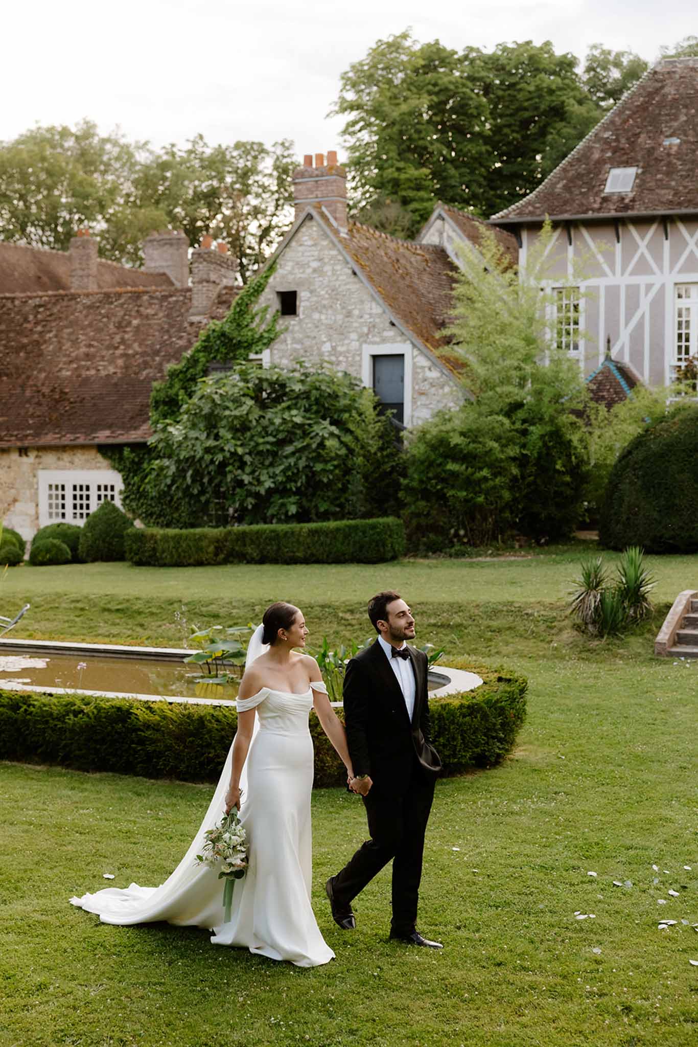 Bride and groom walking hand-in-hand across manicured lawn, historic timber-framed manor buildings in the background.