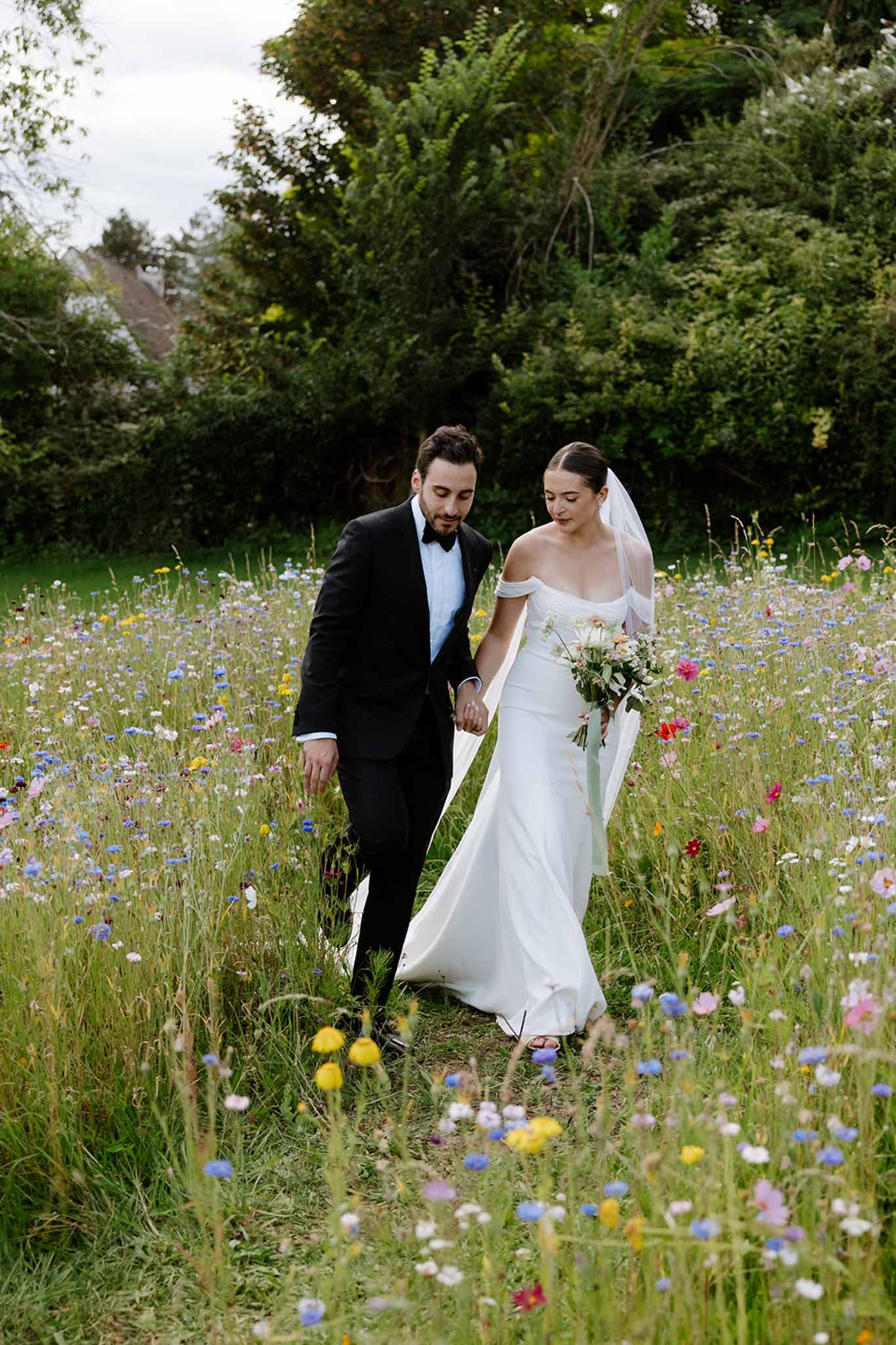 Bride and groom walking hand-in-hand through wildflower meadow with scattered blues, yellows, and pinks, mature trees in background
