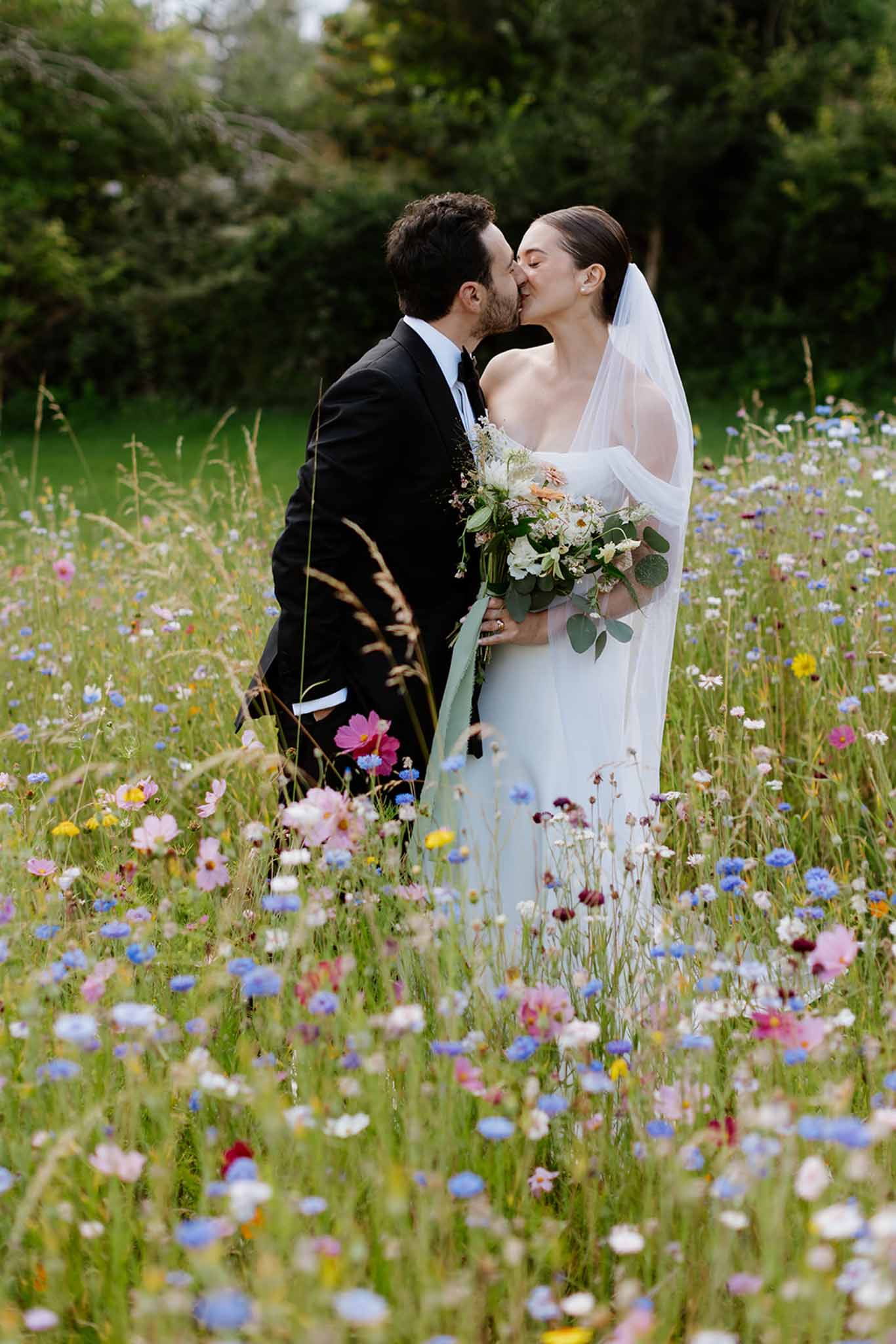 Bride and groom kissing in wildflower meadow with cosmos and cornflowers, bride in ivory gown with veil