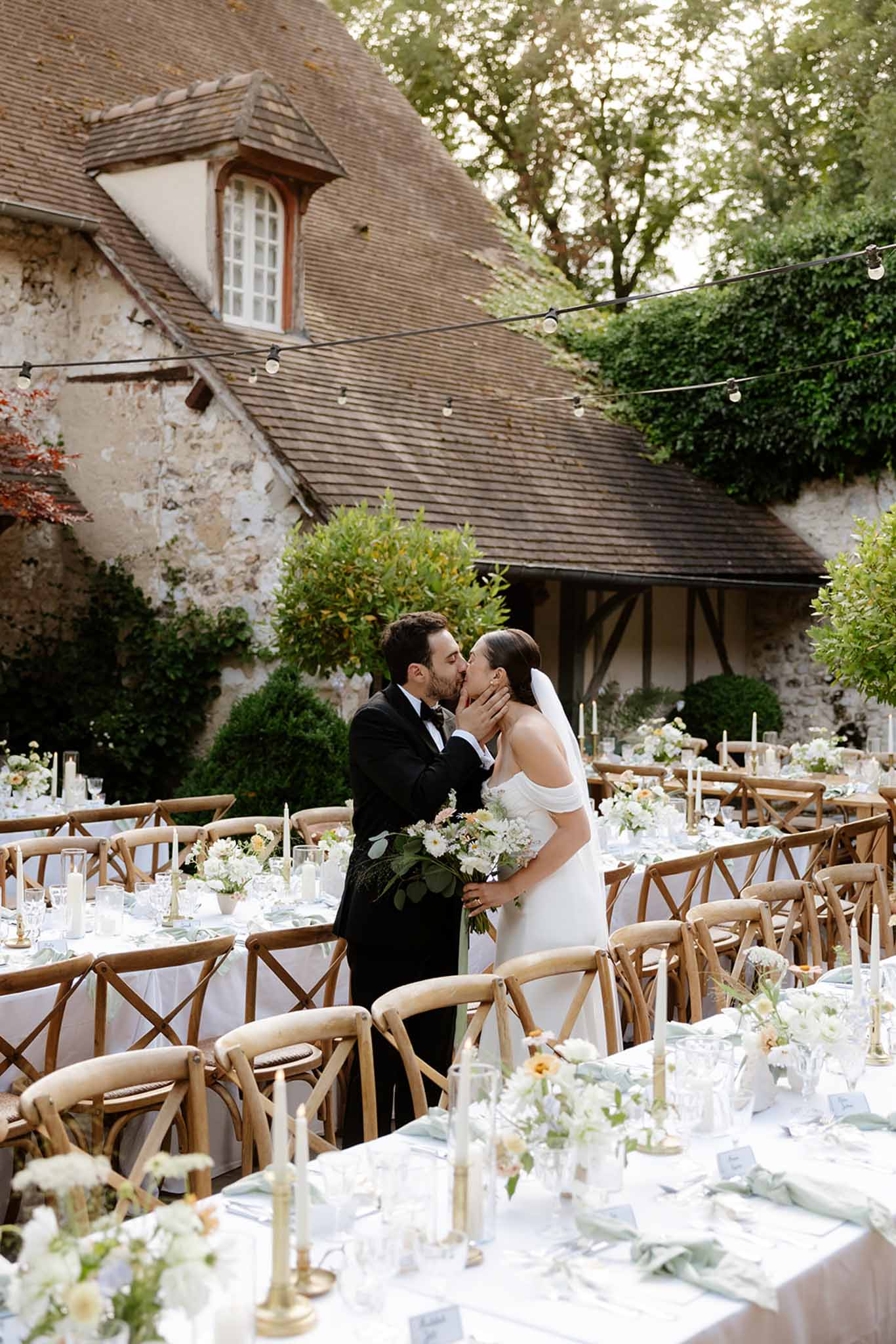 Bride and groom kissing among reception tables in a stone courtyard beside a half-timbered barn with string lights