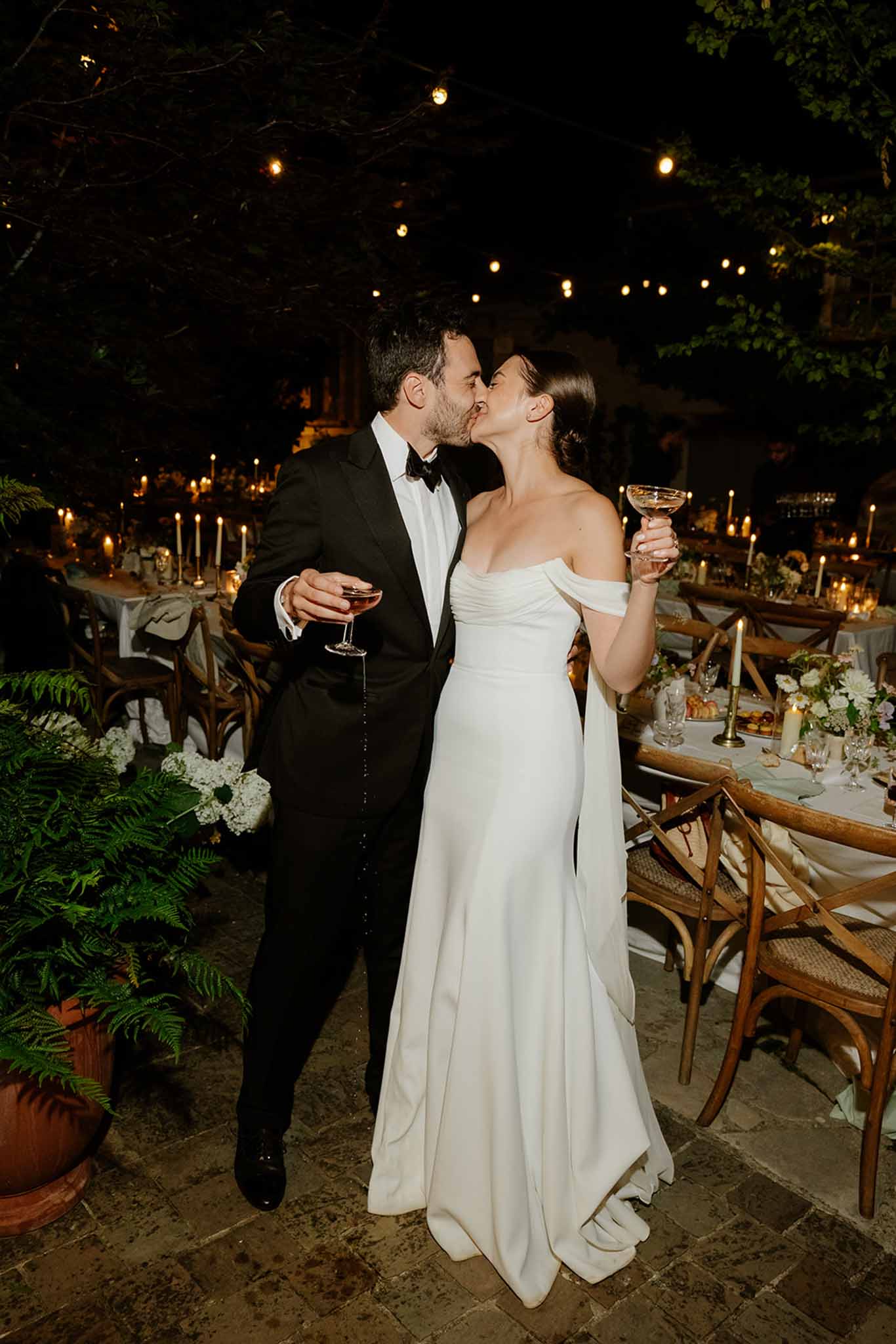 Bride and groom kissing while holding champagne glasses at an outdoor garden reception with bistro lighting