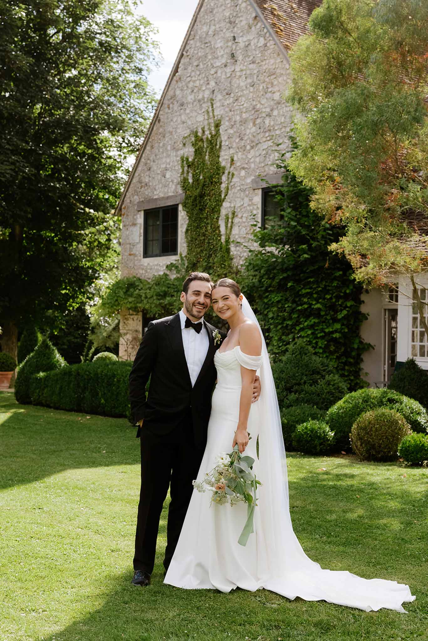 Bride and groom pose on manicured lawn before ivy-covered stone country house with terracotta roof