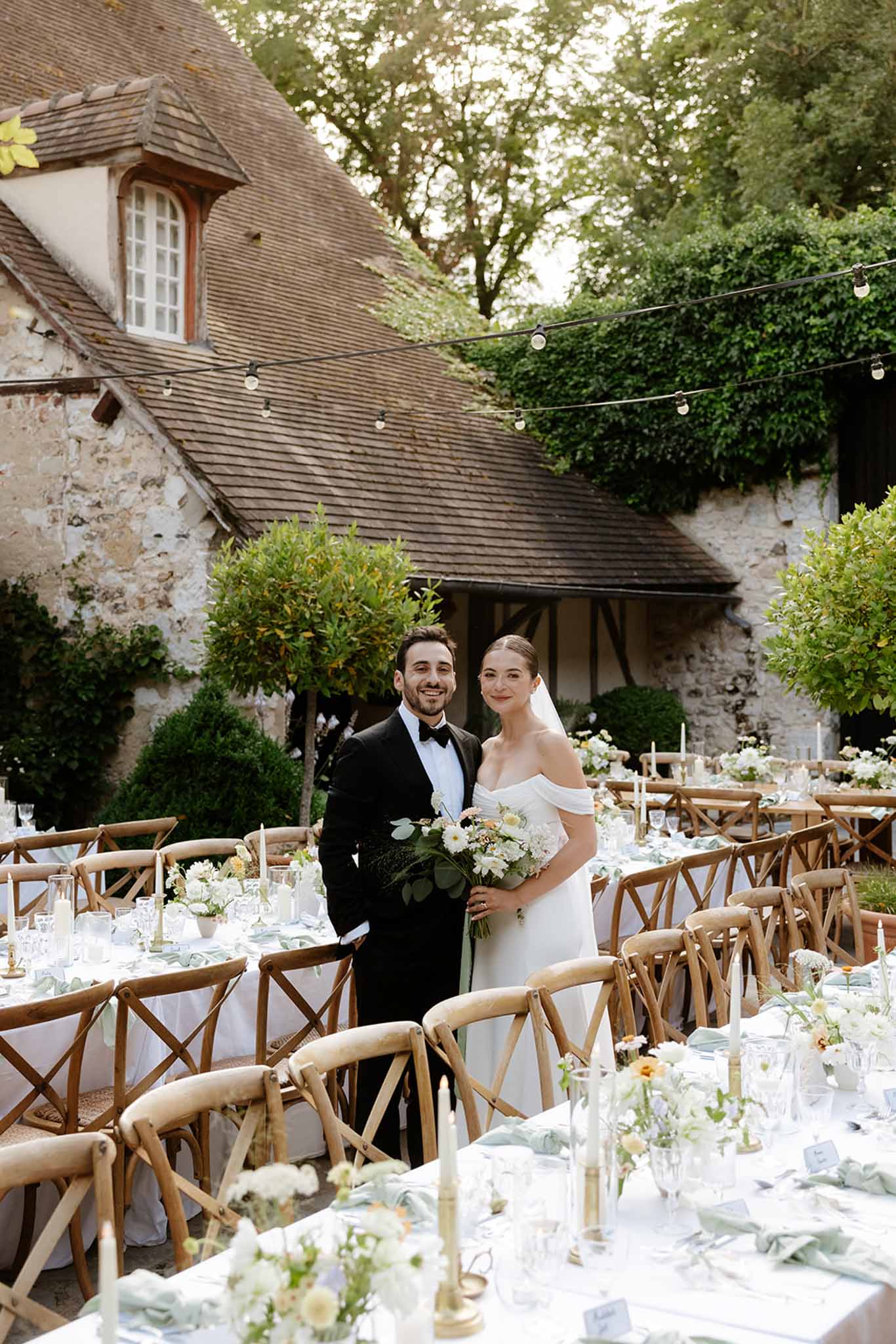 Bride and groom posing in courtyard reception with cross-back chairs, white floral centerpieces, and string lights