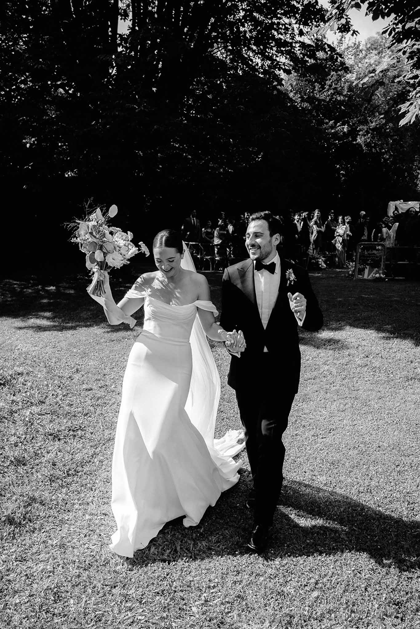 Newlyweds walking through wooded garden in recessional, guests visible in soft focus, black and white photo