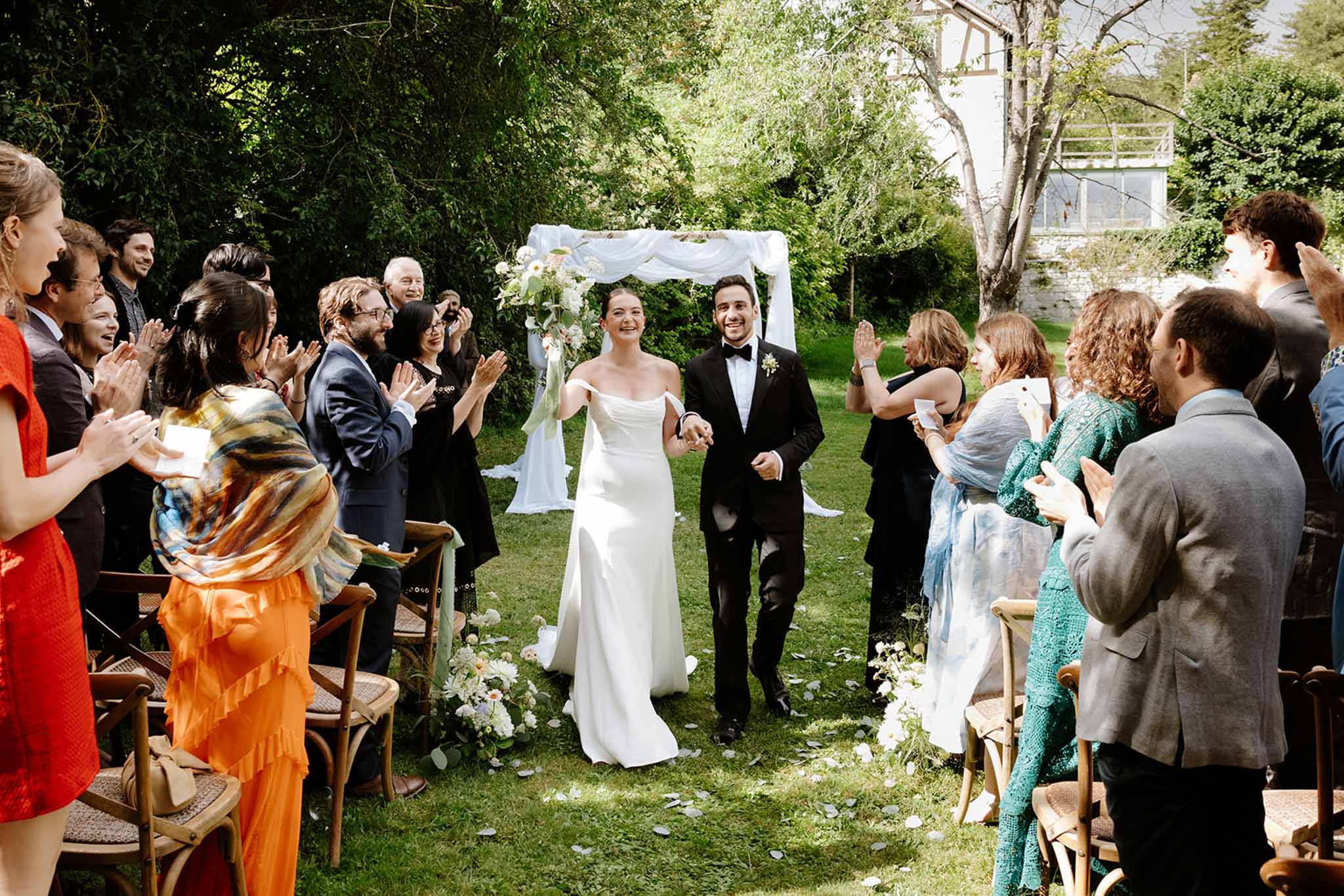 Bride and groom walking aisle recessional while guests applaud at outdoor garden ceremony with stone manor