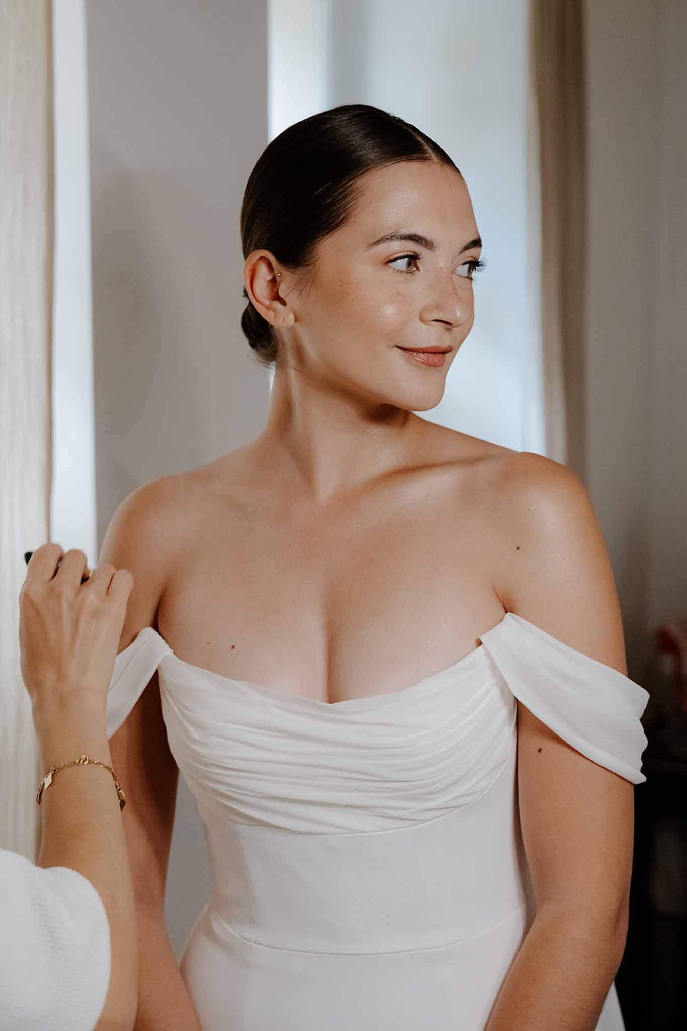 Bride in off-shoulder ivory draped dress with sleek updo photographed against soft white walls during getting ready