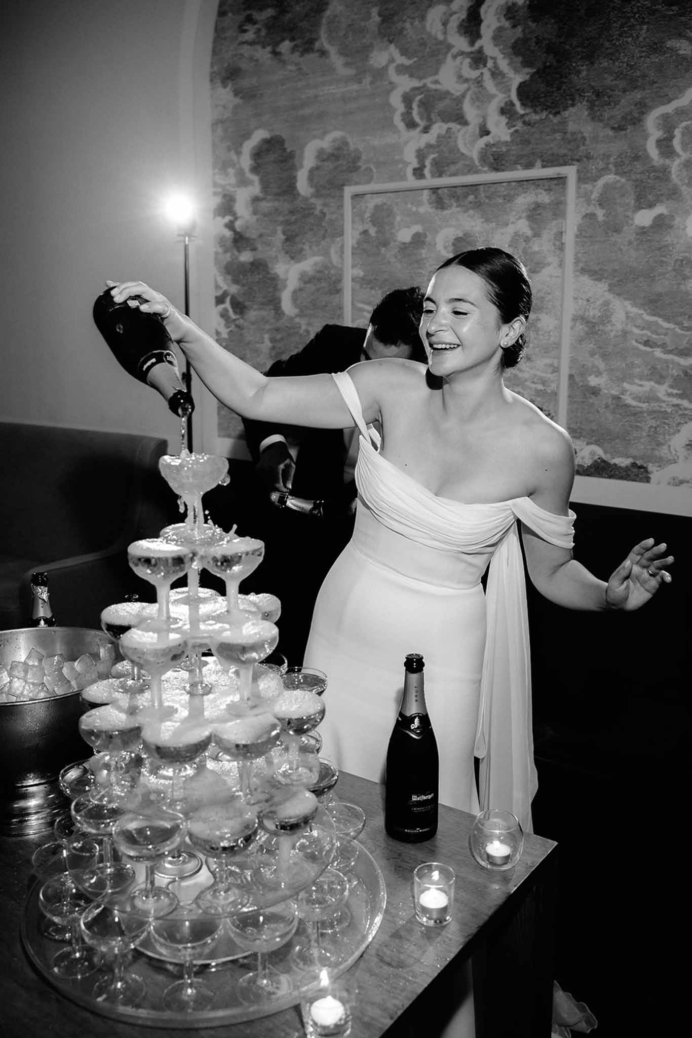 Black-and-white photo of bride smiling while pouring champagne into a coupe glass tower during reception