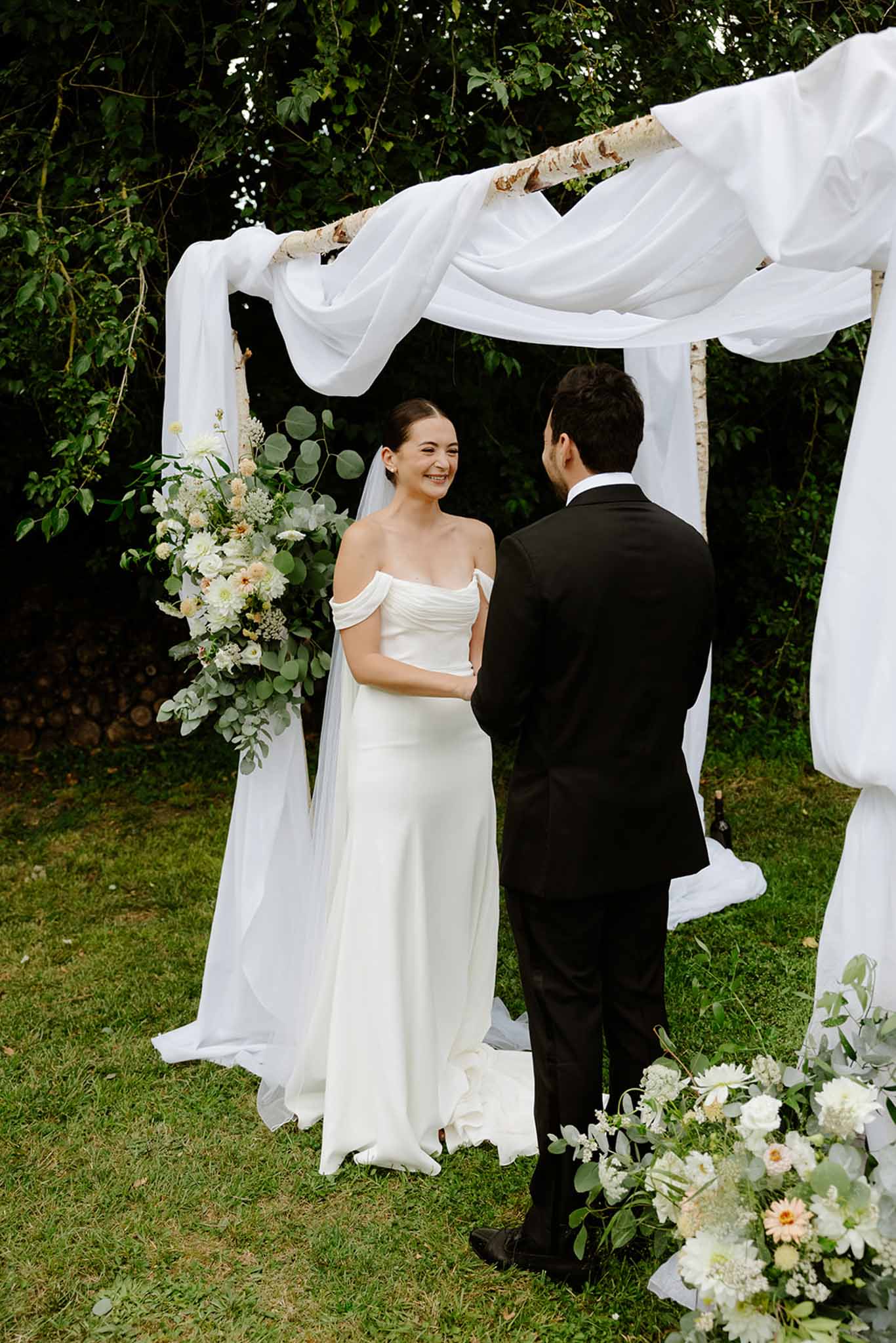 Bride and groom facing each other during outdoor ceremony under white fabric canopy on birch poles in garden