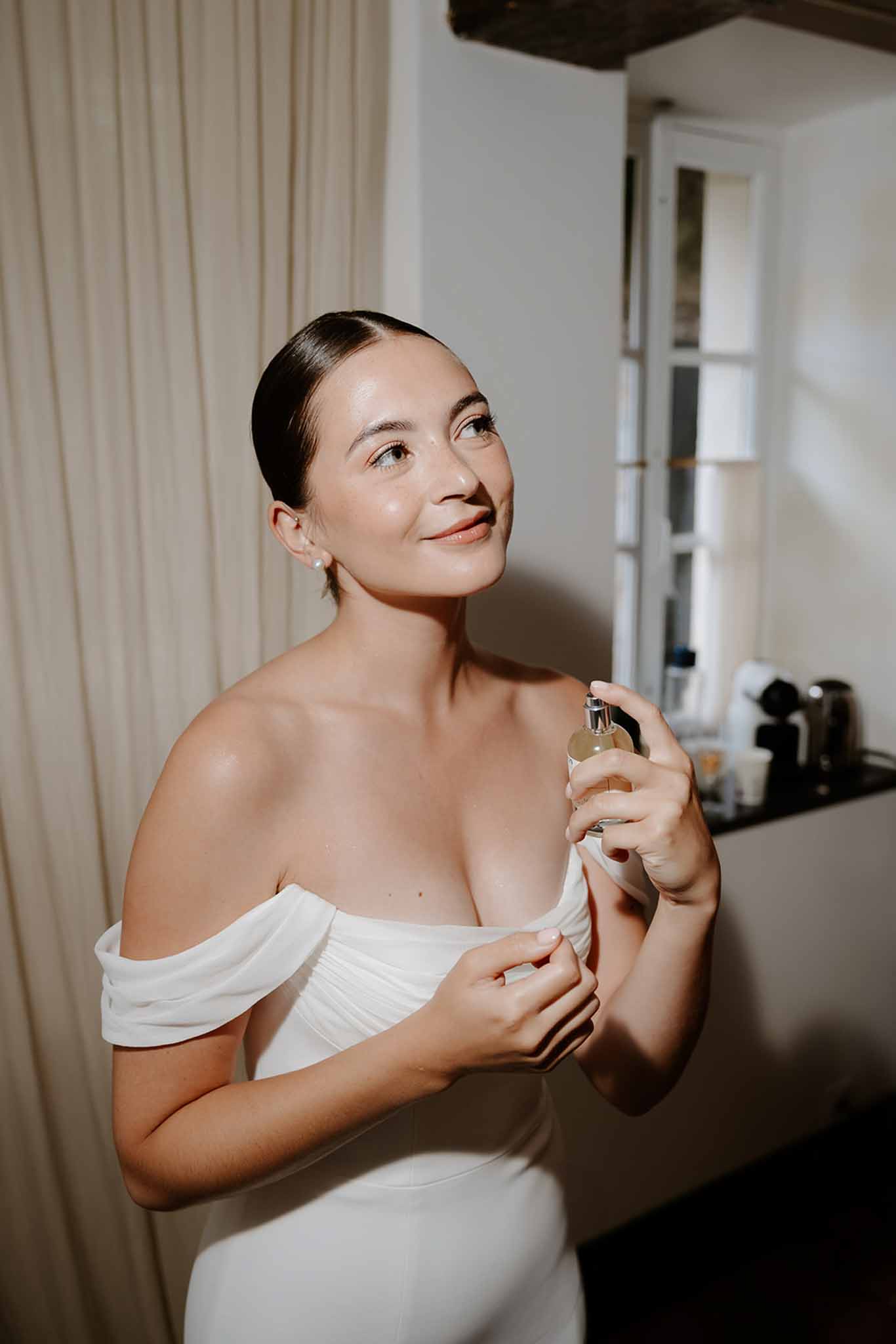 Bride in off-shoulder white dress applying perfume in cream-curtained bridal suite before ceremony