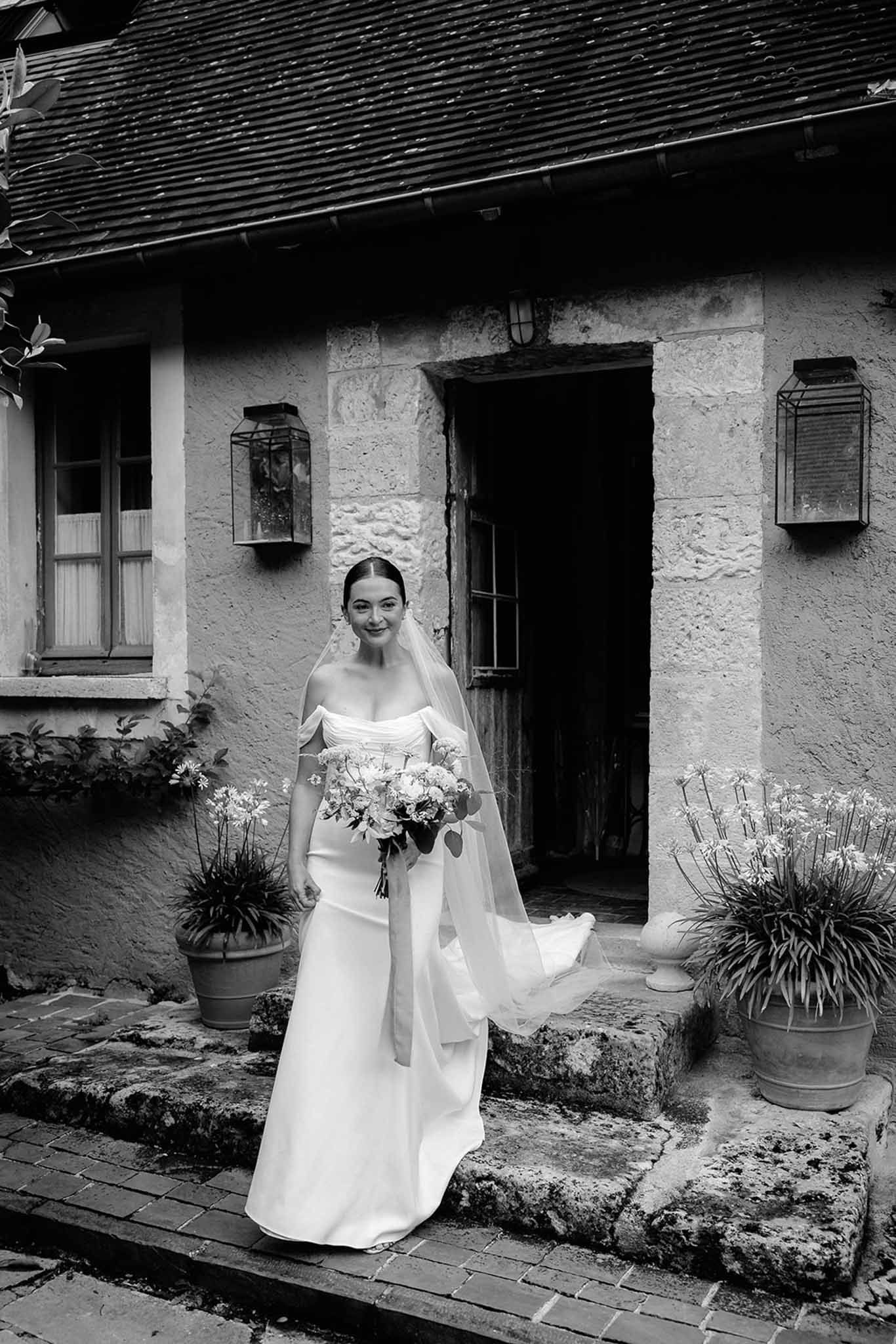 Black-and-white bride in off-shoulder gown standing in courtyard of stone cottage
