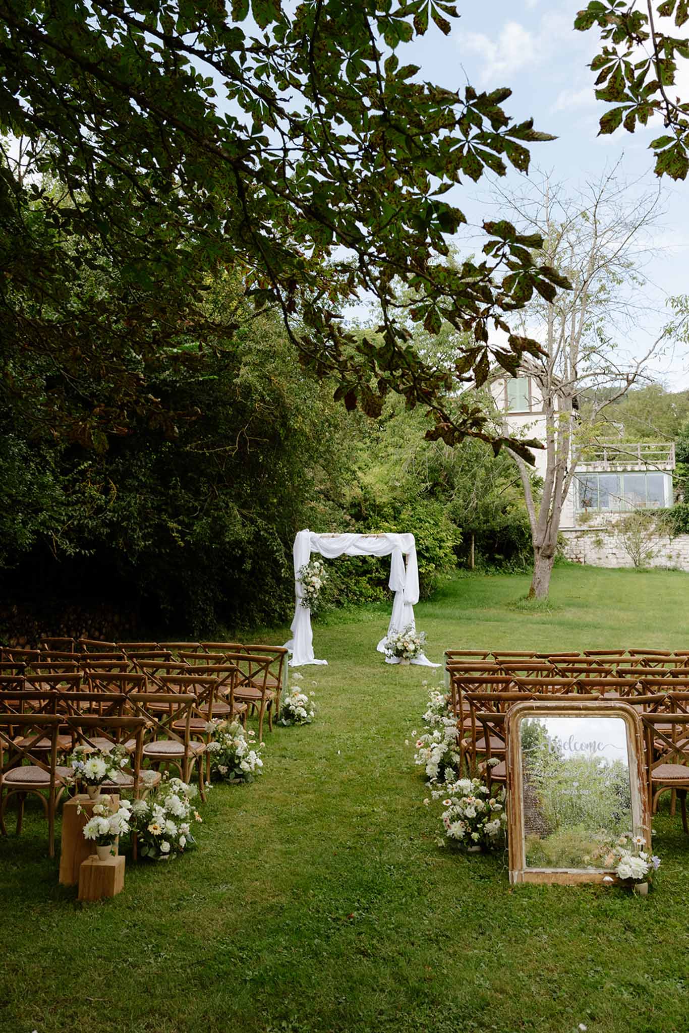 Outdoor garden ceremony setup with wooden cross-back chairs, white floral aisle markers, and draped arch
