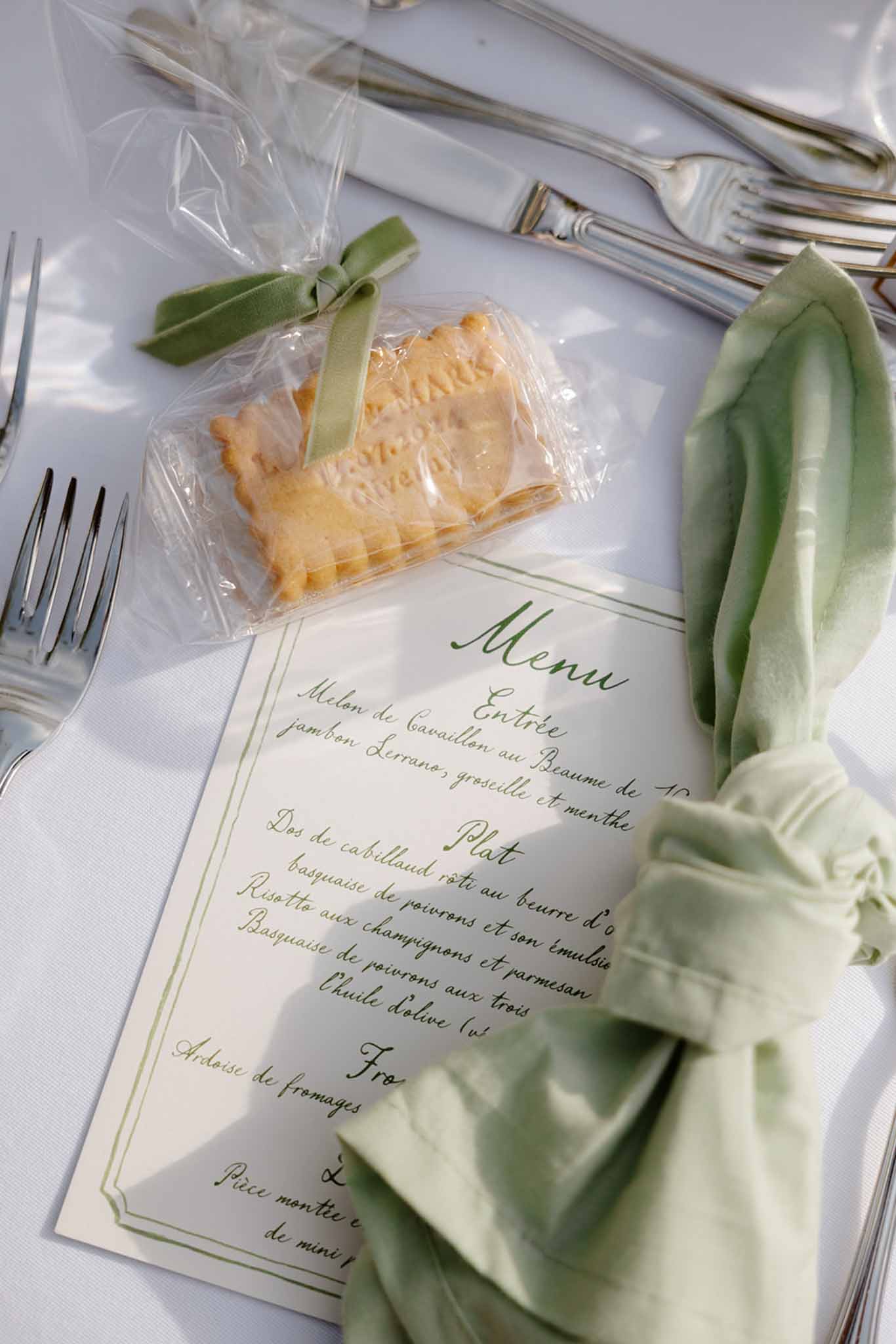 Place setting with handwritten French menu card, sage green napkin, and cellophane-wrapped pastry favour