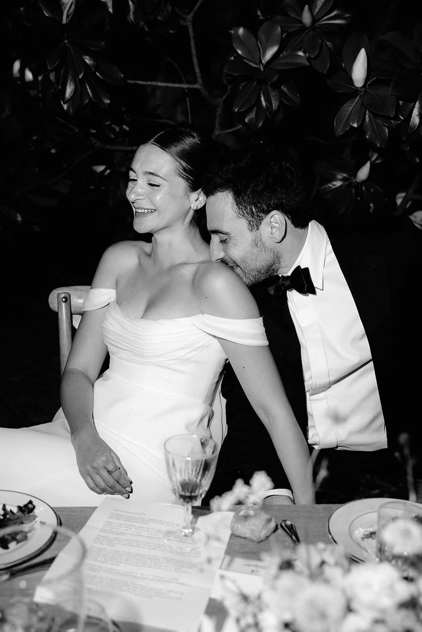 Bride and groom laughing together at outdoor reception table in formal attire, black and white photo