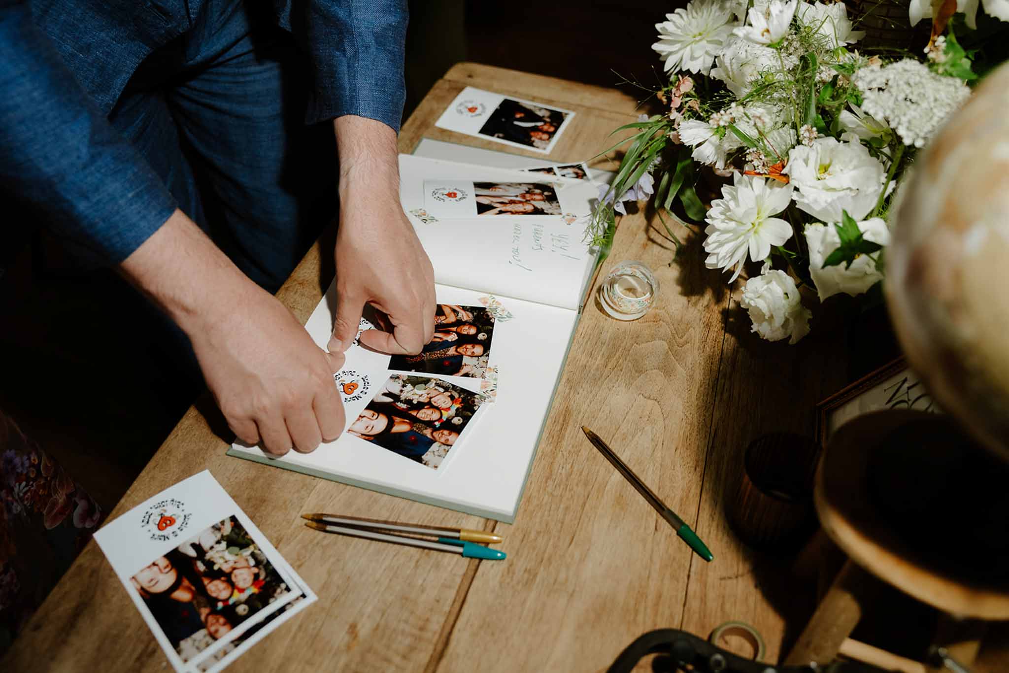 Groom in teal suit examines Polaroid wedding photos in open album beside white daisy and hydrangea arrangement