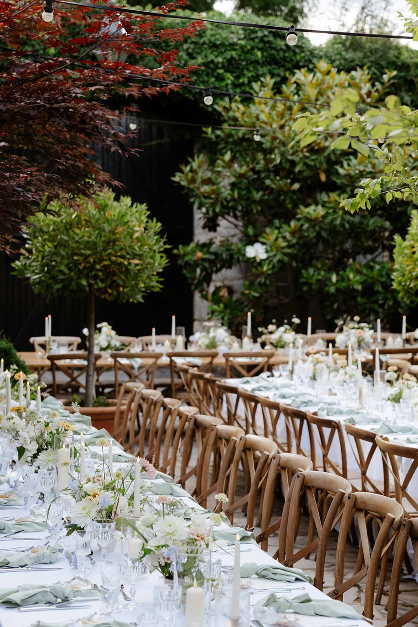 Outdoor reception tables with sage napkins, wooden crossback chairs and white rose centerpieces under string lights