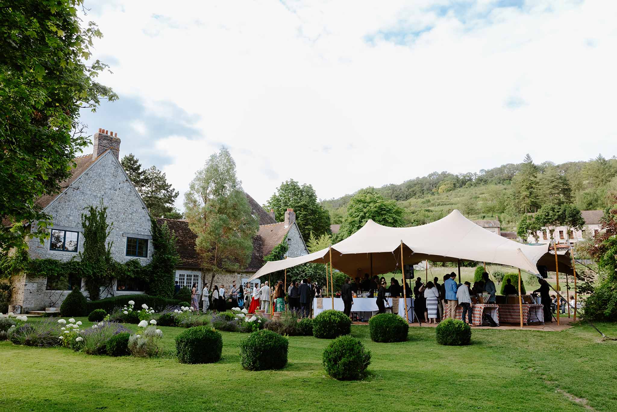 Guests mingling under cream marquee tent on grounds of historic ivy-covered stone property with forested hillside backdrop