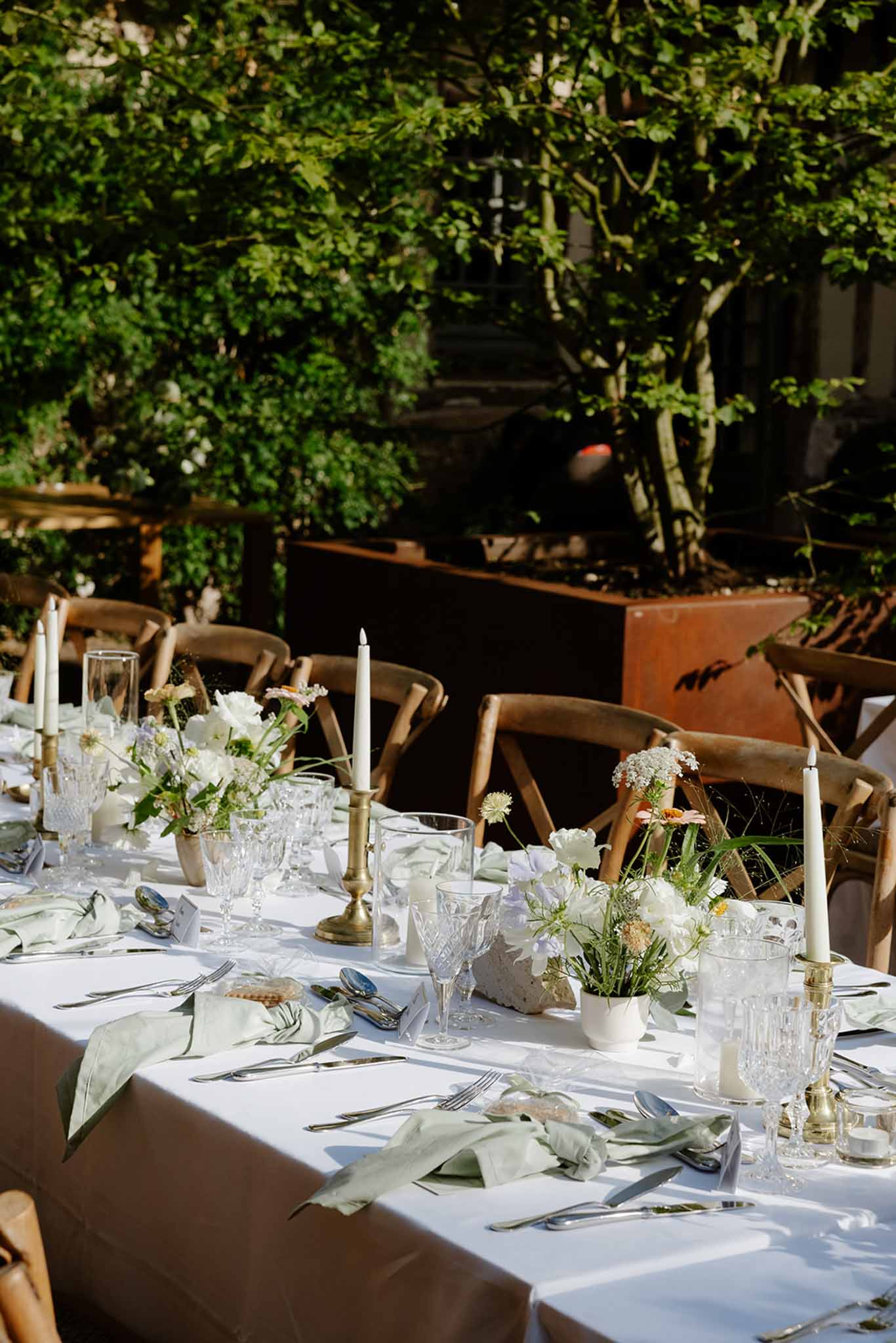 Garden reception table with white ceramic floral vessels, brass candleholders, and climbing ivy overhead