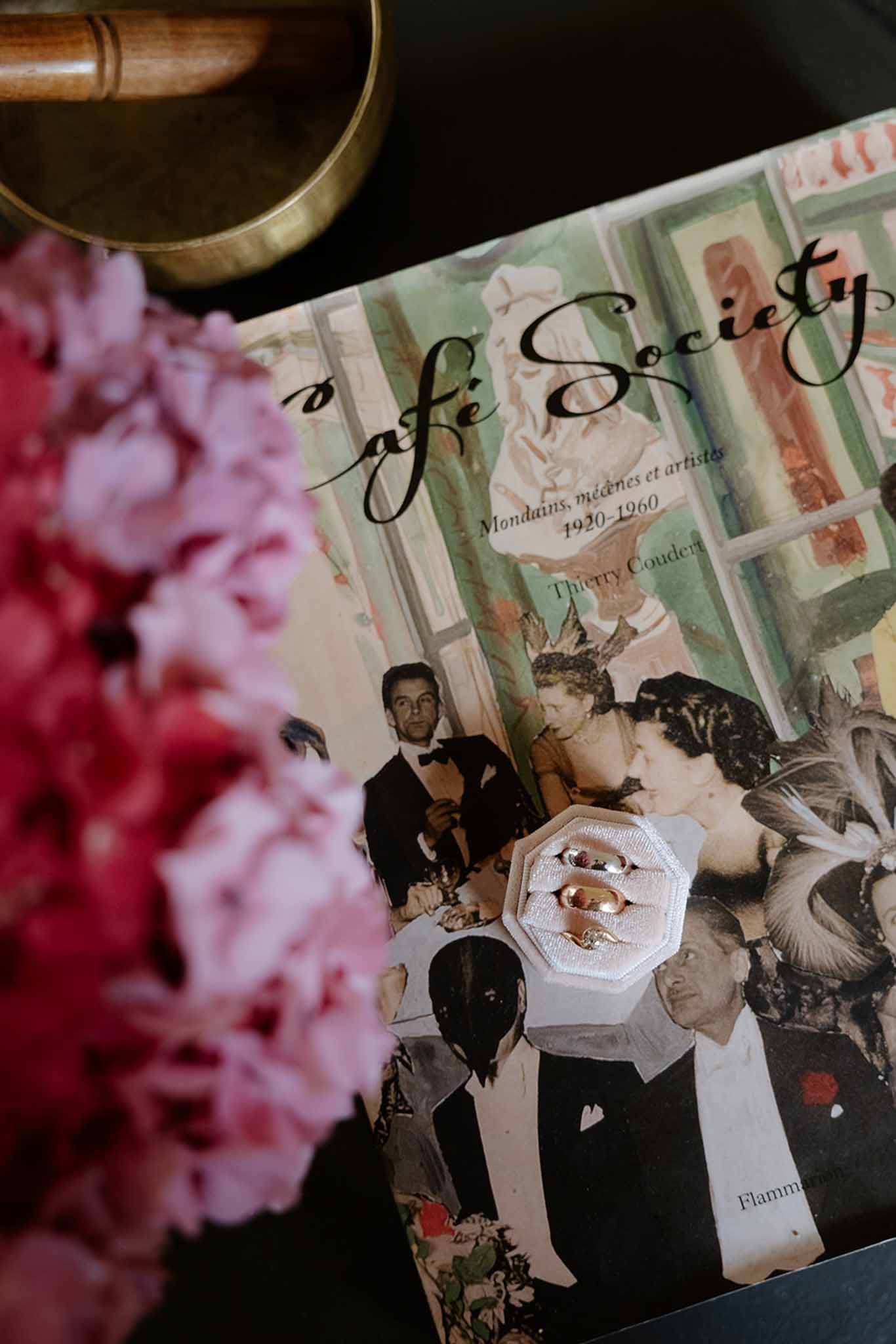 Close-up of two gold wedding bands on hexagonal tile atop vintage Cafe Society book with pink peony