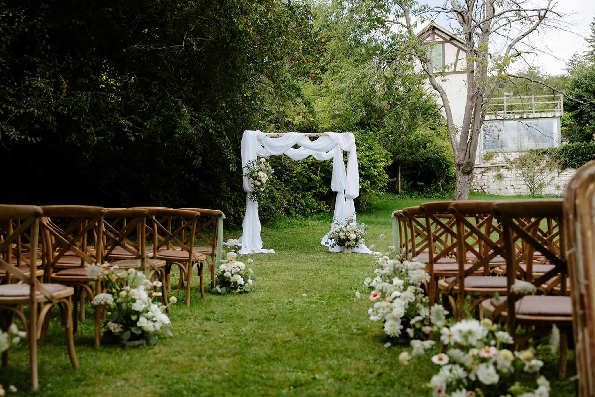 Wedding ceremony at Dime de Giverny with white cloth arch and wooden chairs in outdoor garden setting
