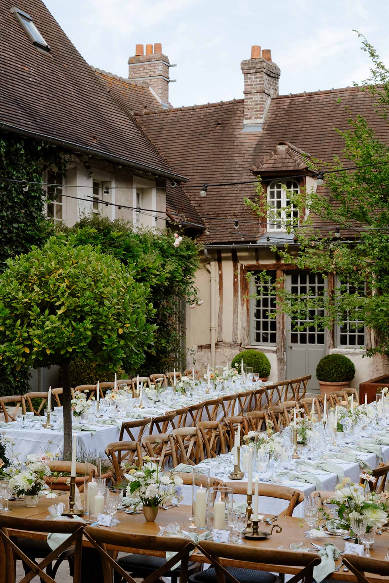 Long wooden reception tables with white linens and flowers set in courtyard beside stone cottage with string lights