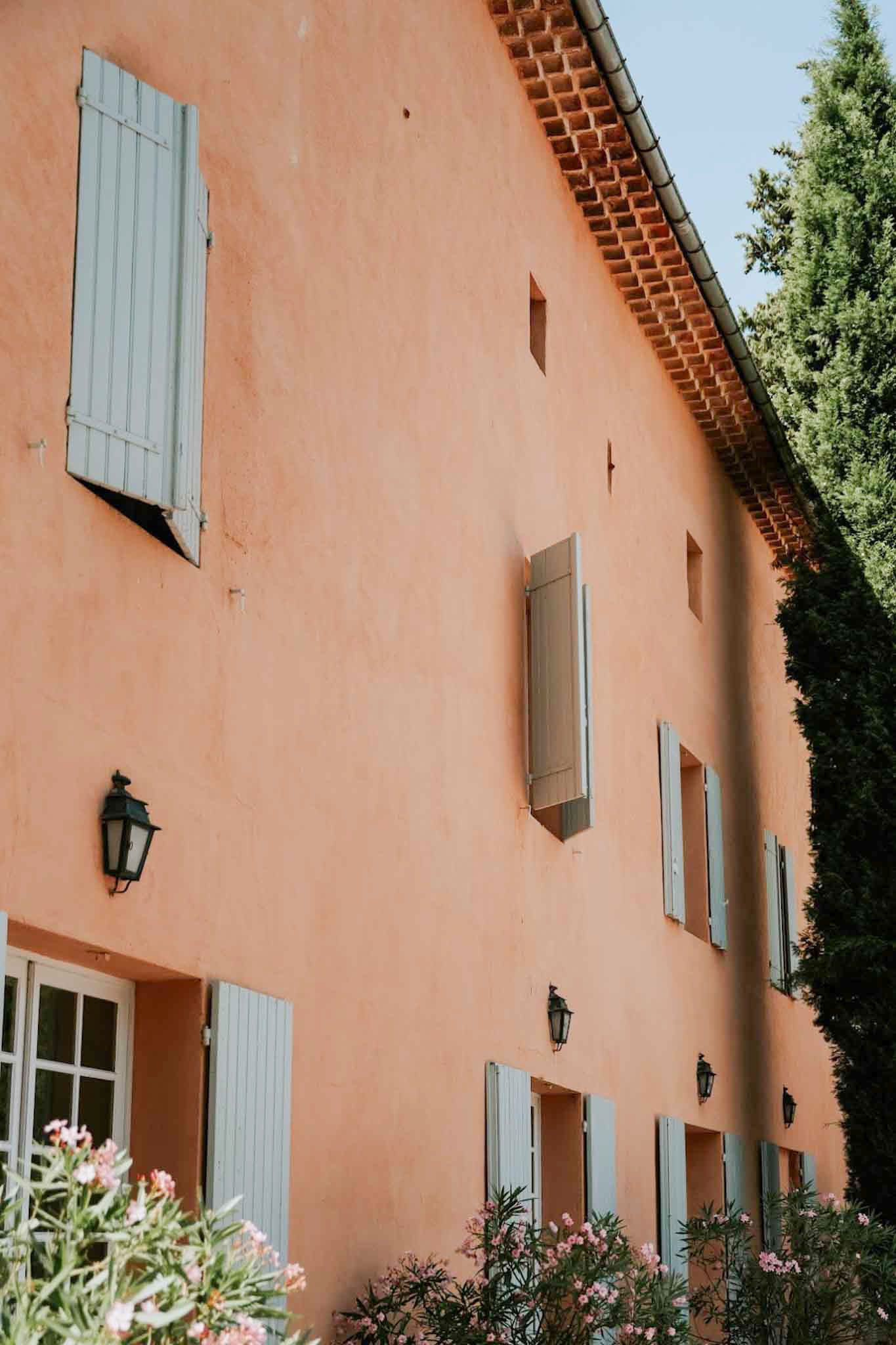 Provencal mas facade with terracotta-orange walls, grey-blue shutters, wrought-iron lanterns, and pink oleander
