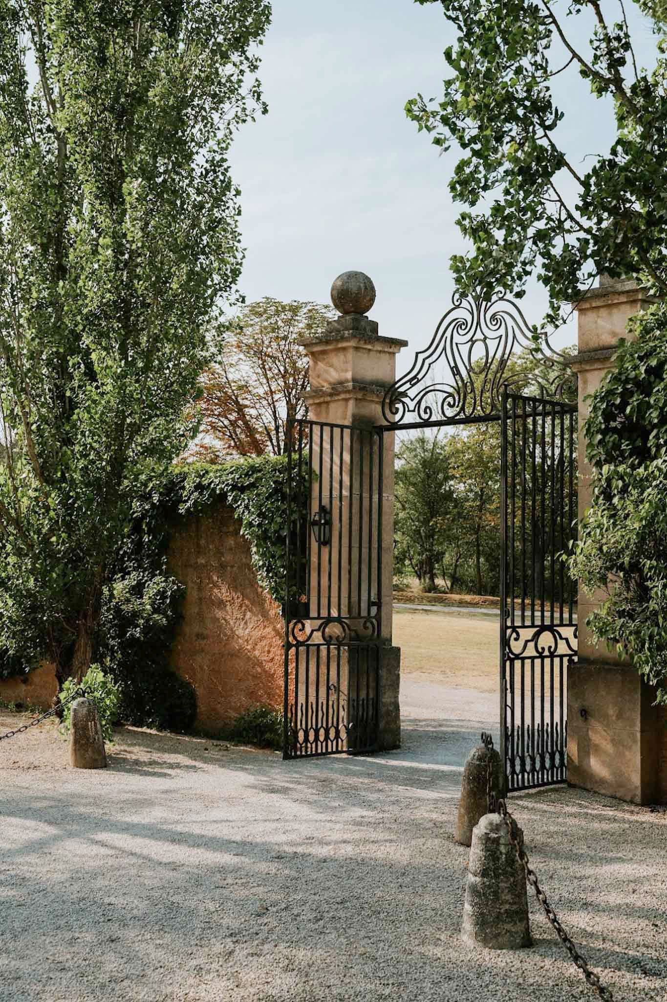 Ornate black wrought-iron gate between stone pillars with ivy-covered walls and gravel driveway entrance