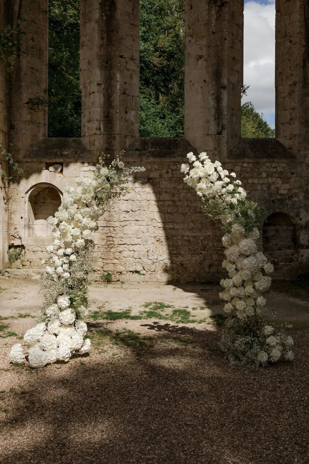 Two tall white rose floral installations flanking open courtyard in historic stone ruins with ivy-covered columns