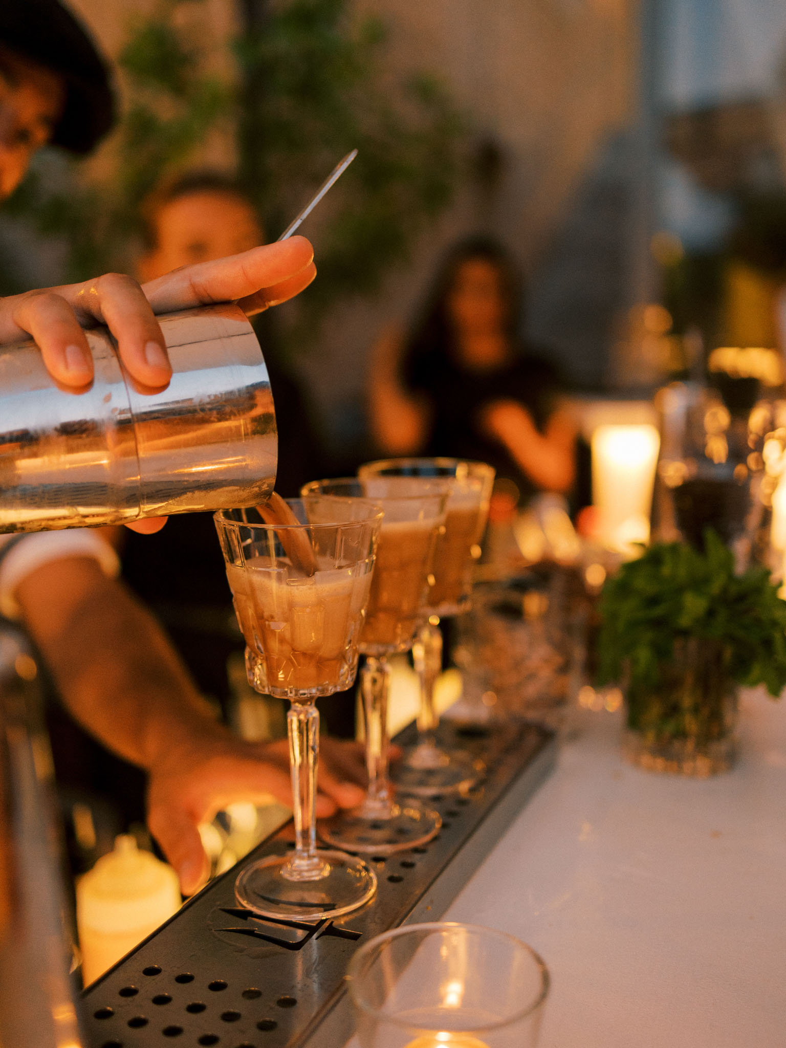 Server pouring amber cocktail into coupe glasses at outdoor bar during golden hour