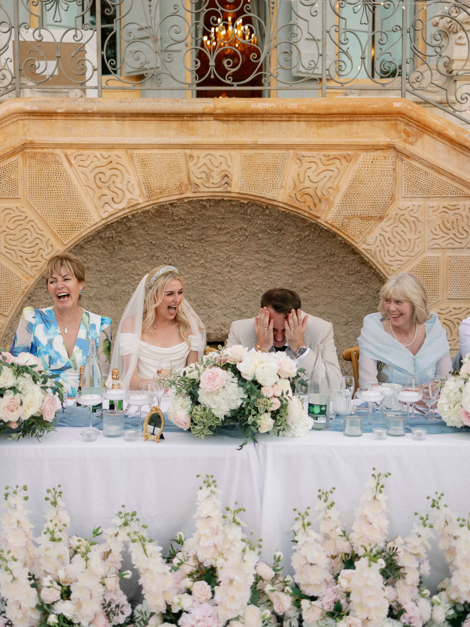 Bride and groom at reception top table with family in ornate stone courtyard with pink and white roses