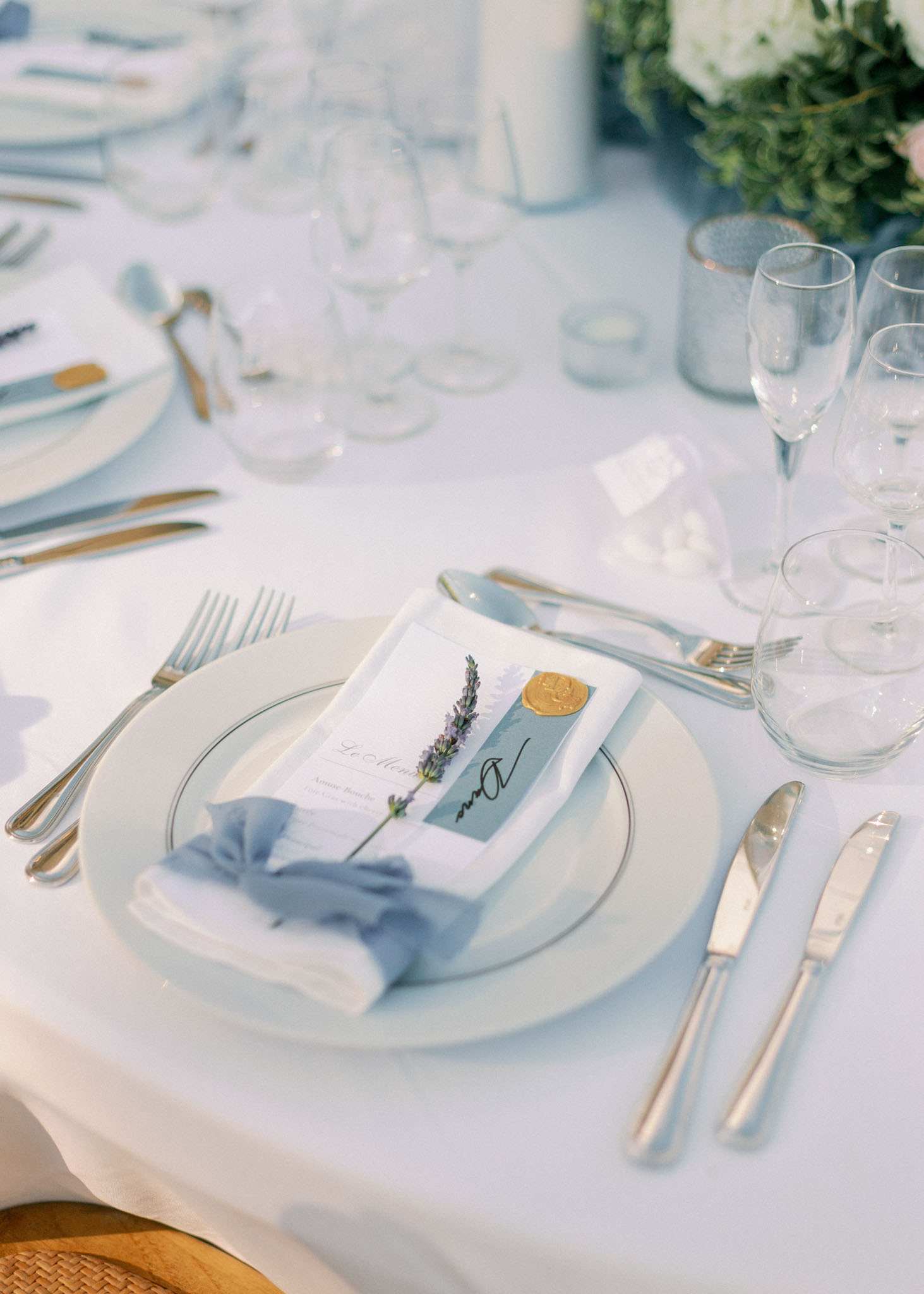 Close-up place setting with light blue napkin, lavender sprig, gold wax seal place card, and silver flatware