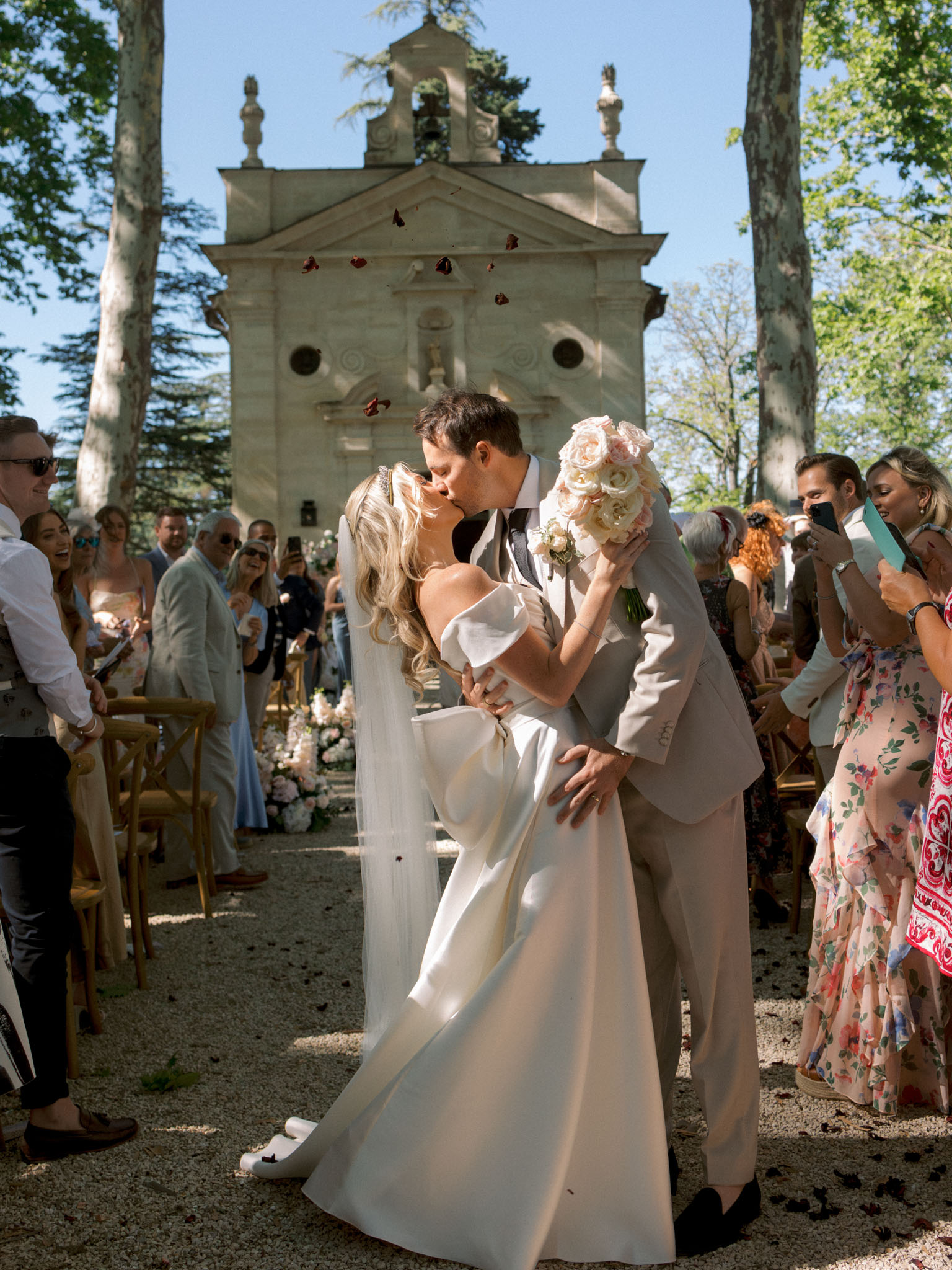 Bride and groom kissing during recessional with guests tossing rose petals at stone chapel