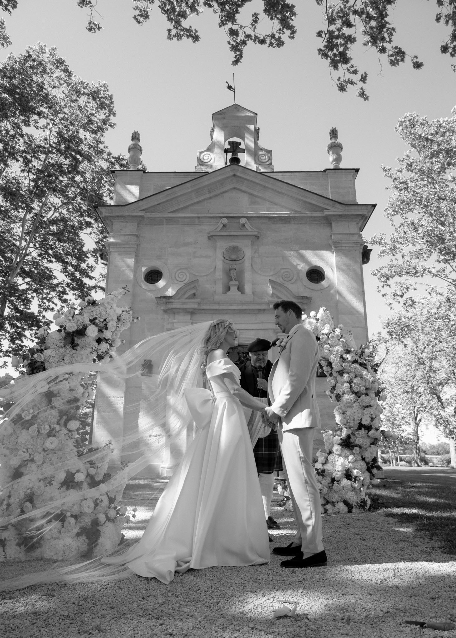 Black-and-white wide shot of wedding ceremony in front of stone chapel with bell tower and white floral arrangements
