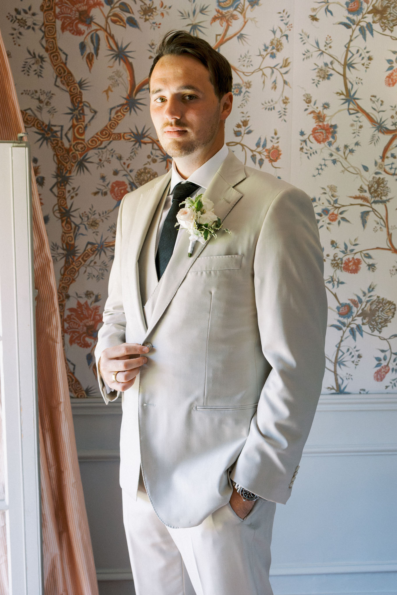 Groom in cream suit with charcoal shirt and black tie, white boutonniere pinned to lapel, posed against botanical-patterned wallpaper.