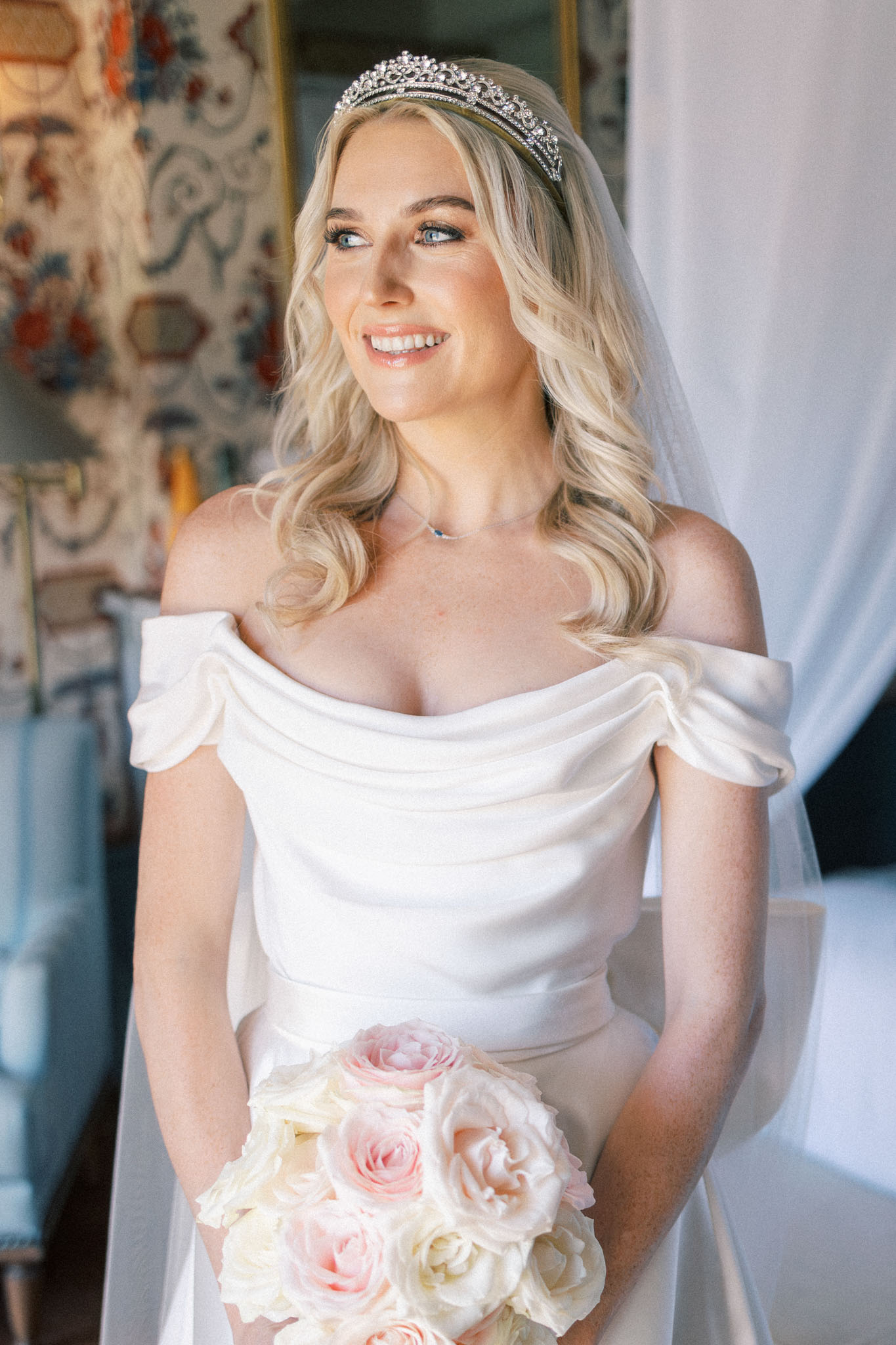 Bride in off-shoulder white dress with crystal tiara and veil holding bouquet of ivory and pink roses