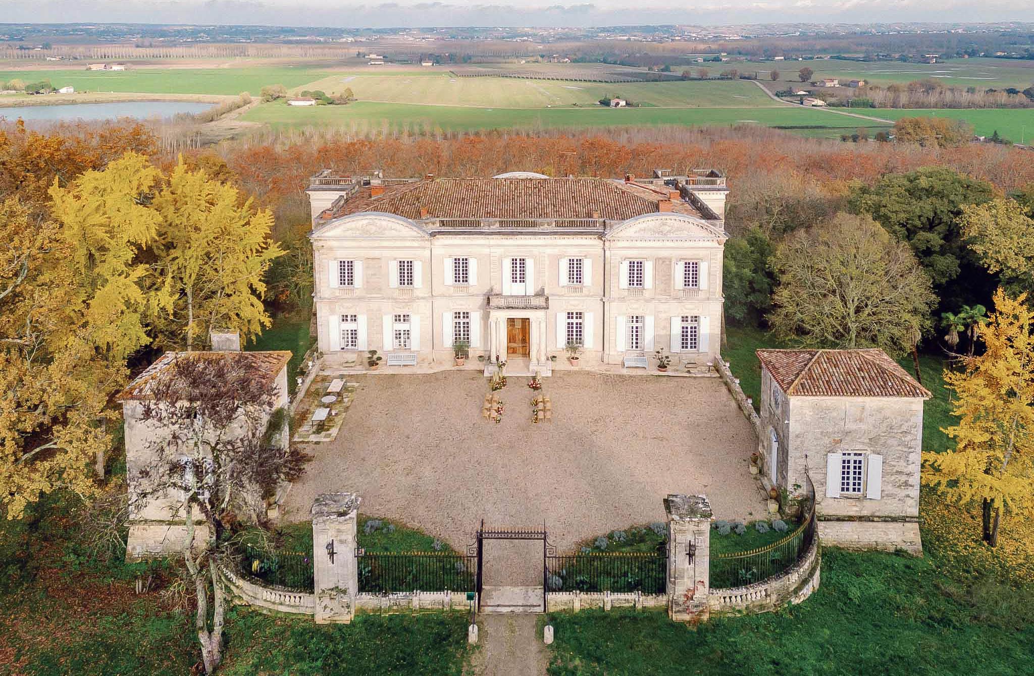 Aerial shot of a symmetrical cream neoclassical château with terracotta roofs, gravel courtyard and autumn foliage surroundings.