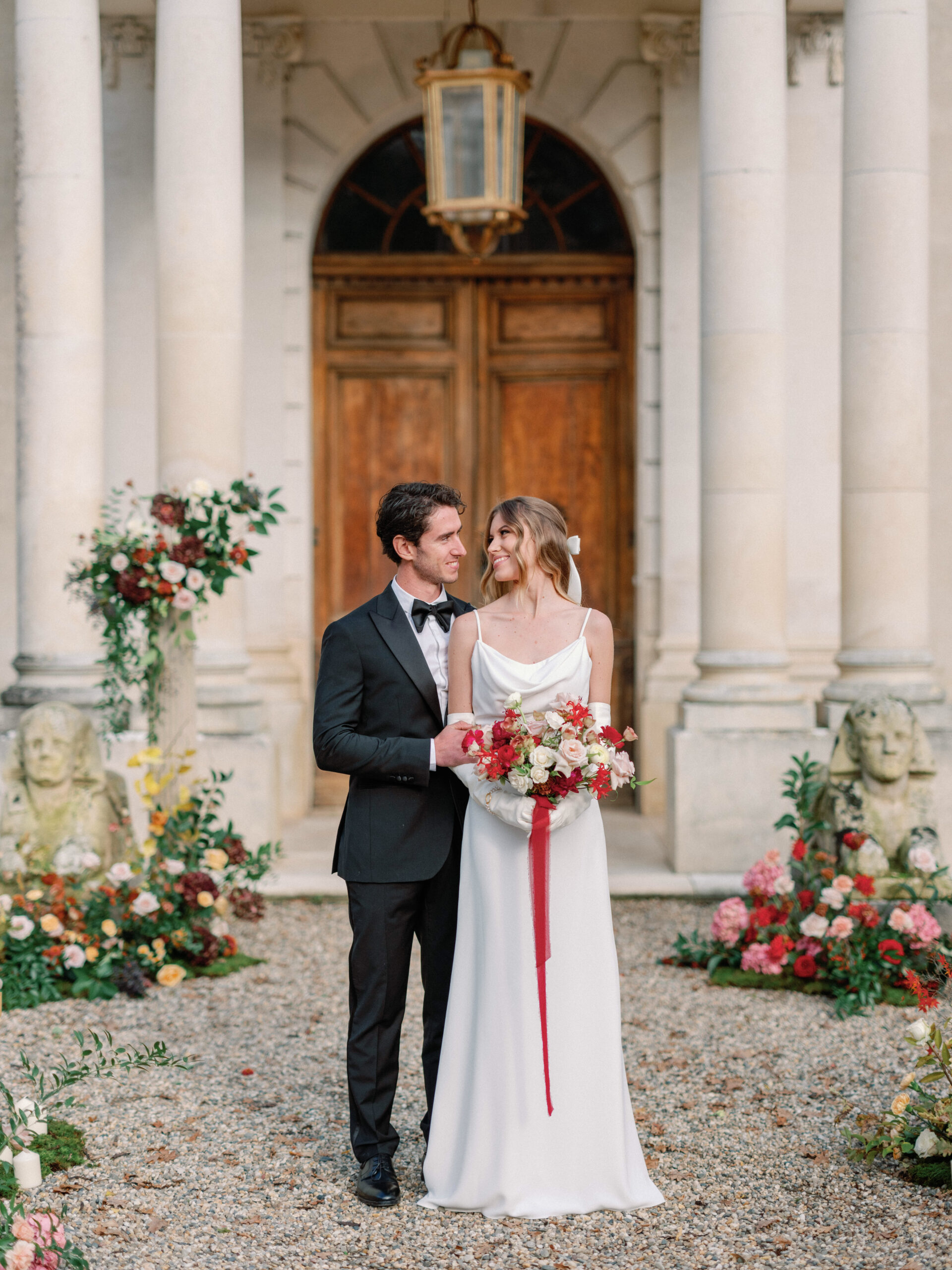 Couple at chateau entrance with jewel-toned floral busts, bride in satin slip dress holding crimson-ribbon bouquet